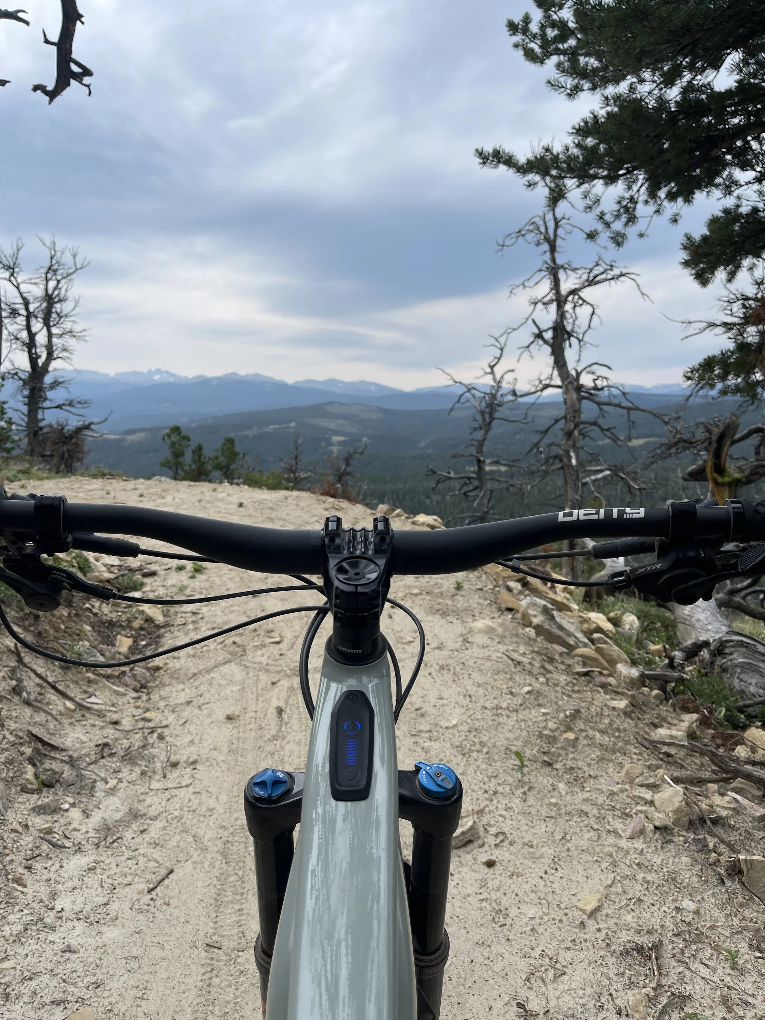 A mountain bike viewed from the rider's perspective on a dirt trail. The trail has dirt and small rocks, with a scenic landscape of mountains, trees, and cloudy sky in the distance.