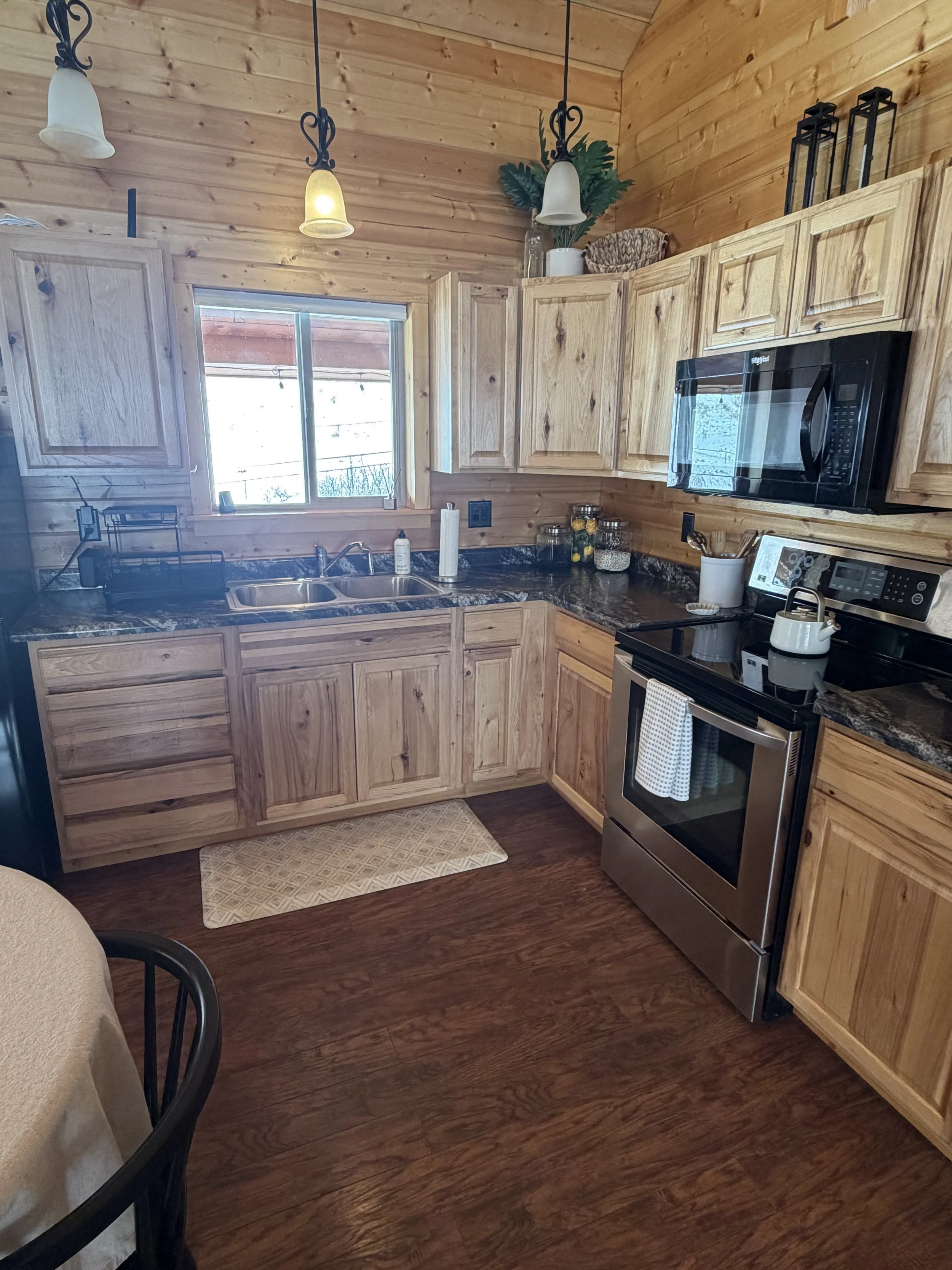A cozy kitchen with wooden cabinets, a dark countertop, stainless steel oven, microwave, and wood-paneled walls. There is a window above the sink, decorative jars, potted plant, and pendant lighting.