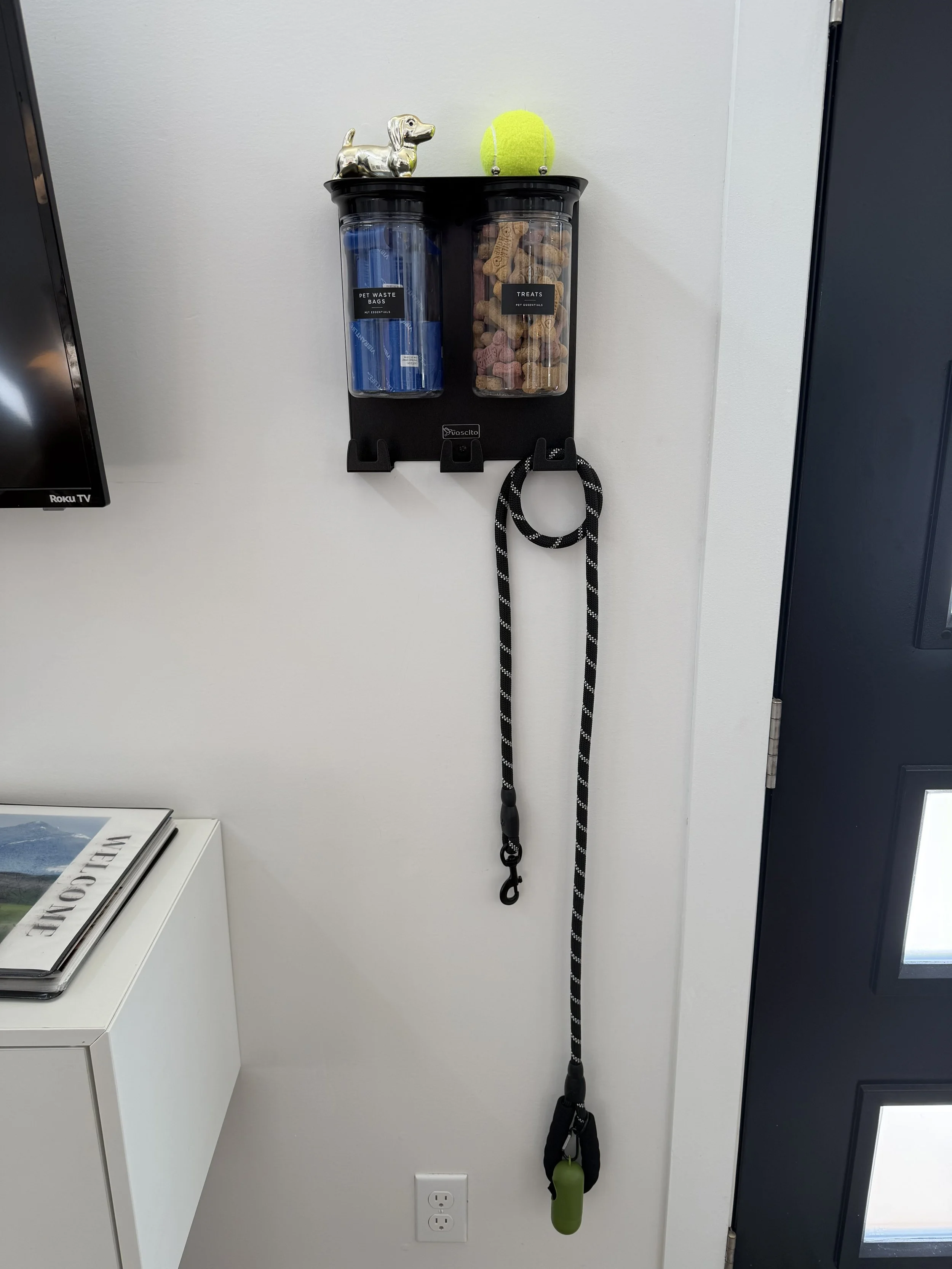 Wall-mounted organizer with two clear containers holding pet waste bags and dog treats, a leash hanging down, a small dog figurine, and a tennis ball on top.