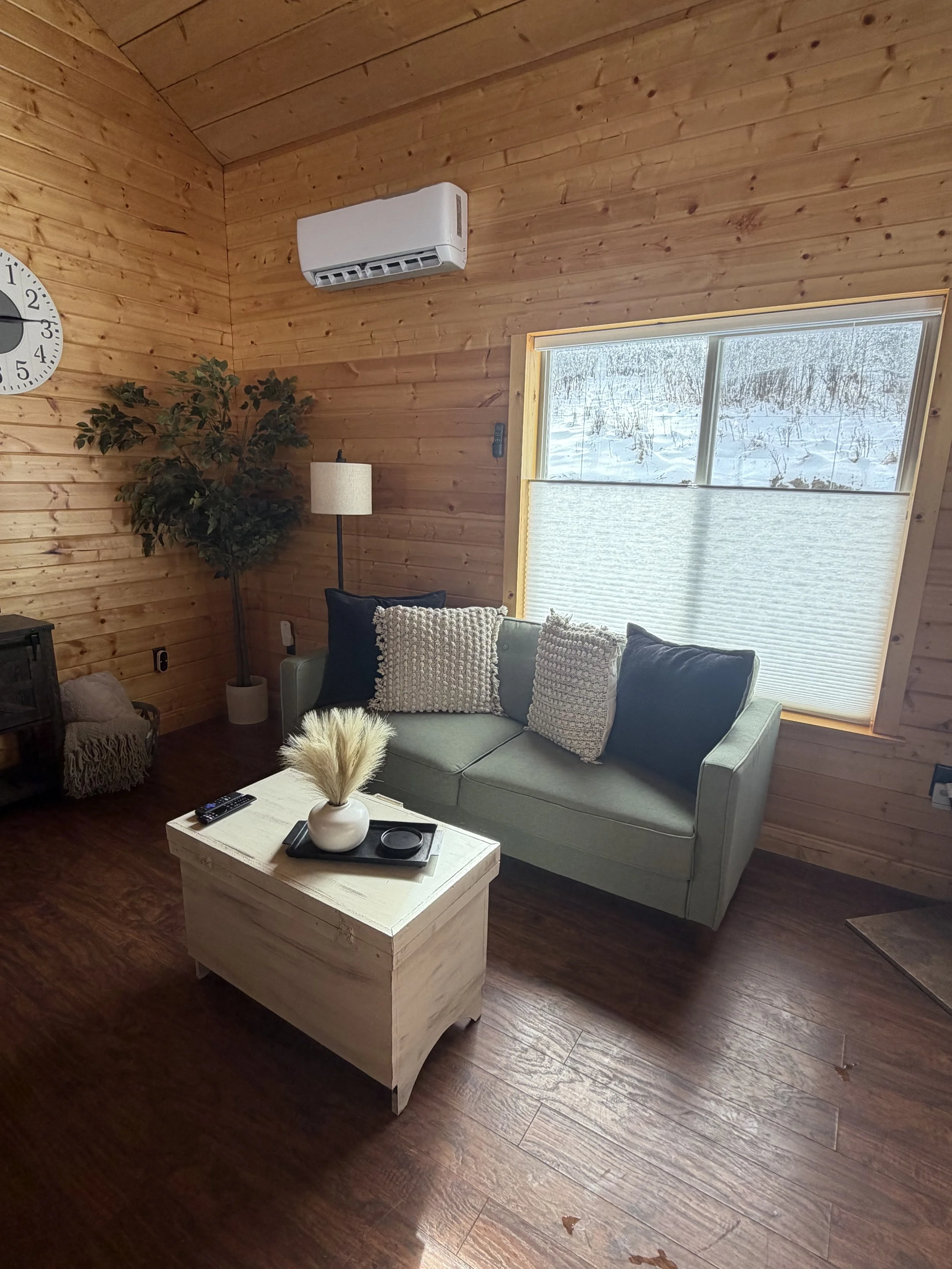 Living room with wooden walls, a window with a view of snow outside, a light green sofa with decorative pillows, a black side table, a potted plant, a floor lamp, a white coffee table with decorative items, and a wall clock.