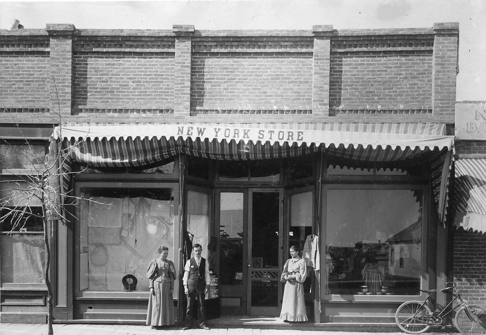 Historic black-and-white photo of the New York Store storefront in Sheridan, with four people and a bicycle out front.