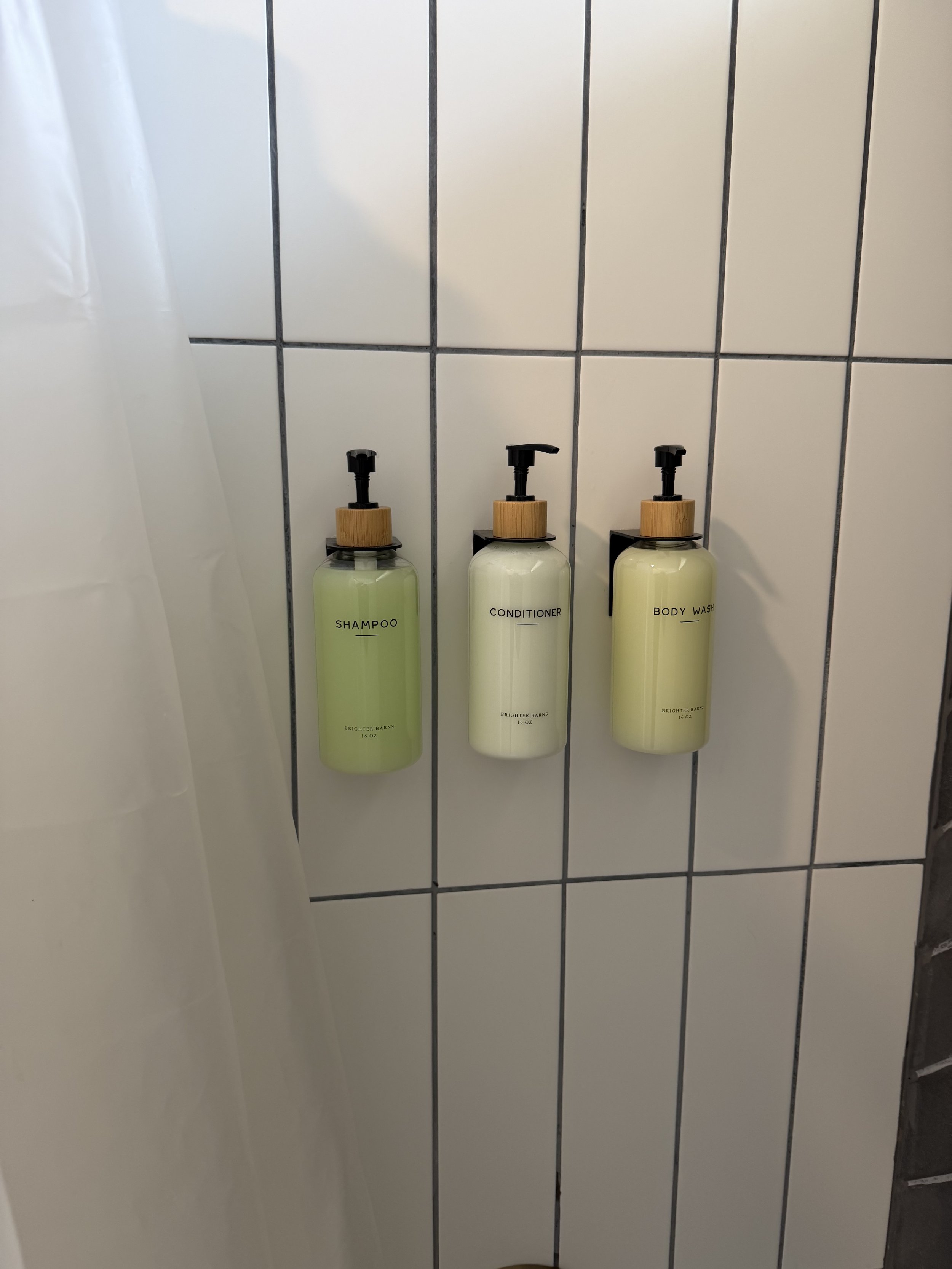 Three wall-mounted bottles of shampoo, conditioner, and body wash in a bathroom with white tiled wall.