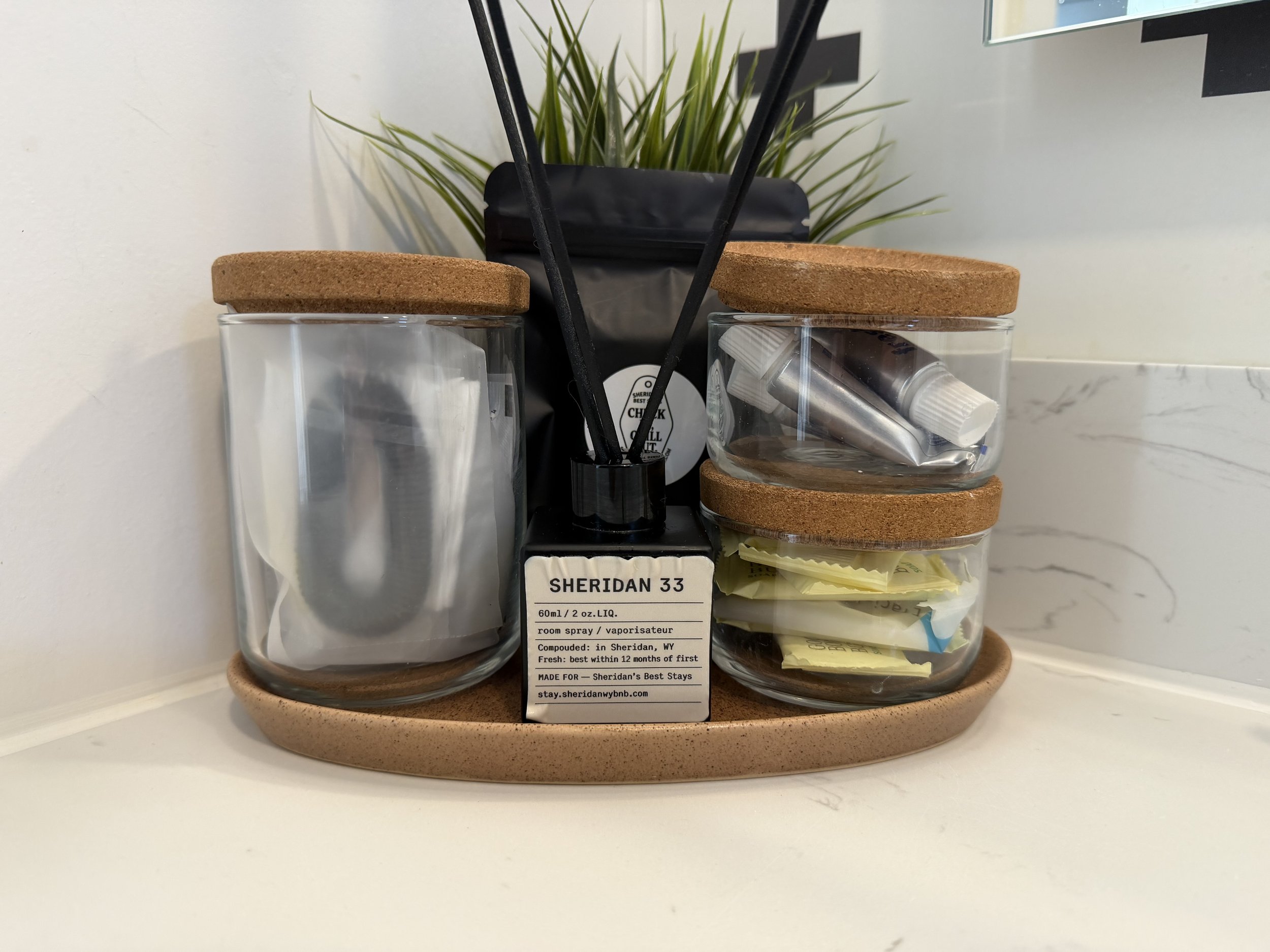 Organized countertop with three glass jars with cork lids, containing utensils, toiletries, and sugar packets, with a small black diffuser in the center, a black bag behind, and a plant in the background.