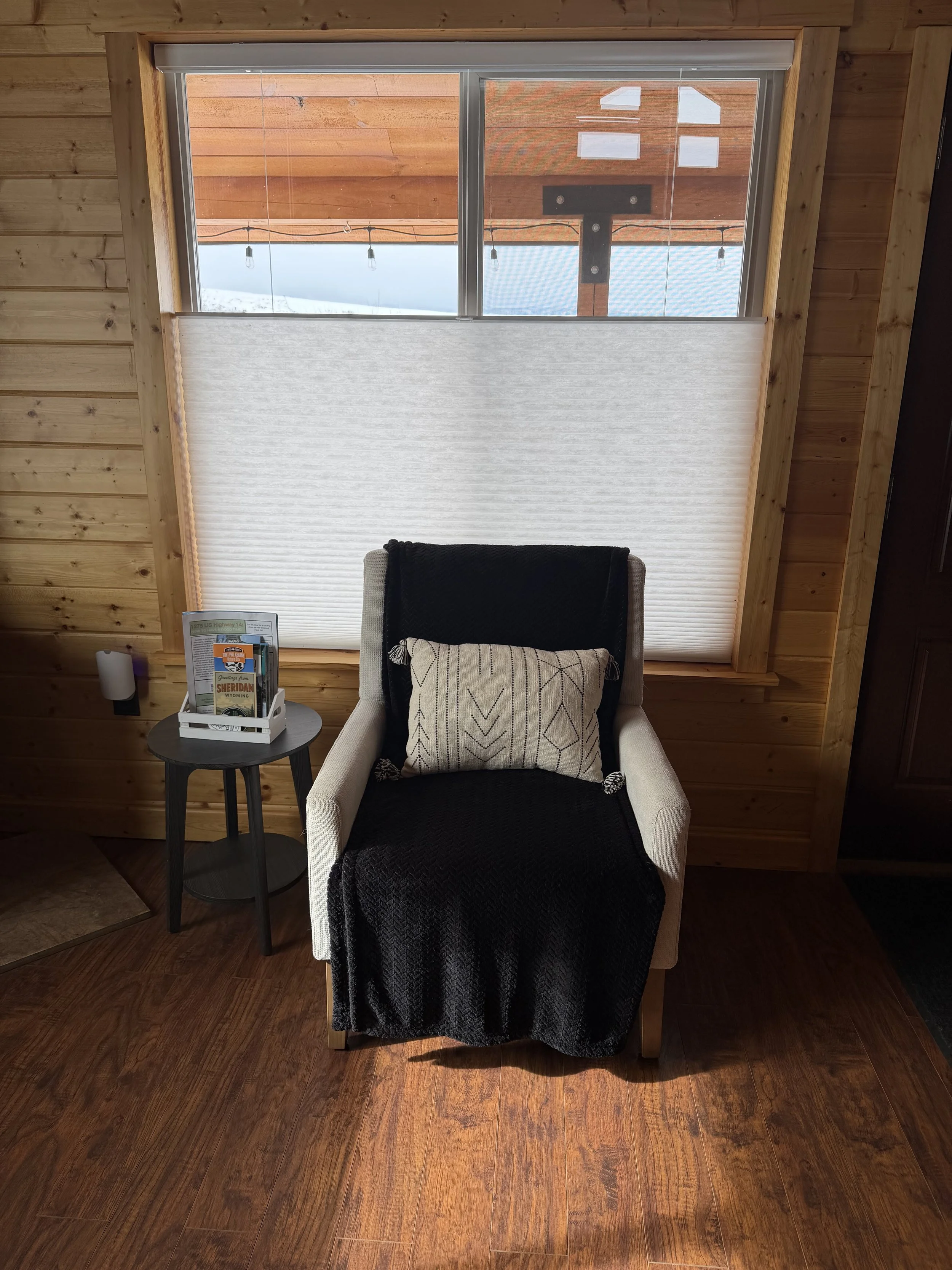 Cozy corner inside a wooden cabin with a beige armchair draped with a black blanket and decorated with a patterned throw pillow. A small round side table holds a magazine rack. Behind, a window with a partly closed white blind shows an outdoor pergola with string lights.