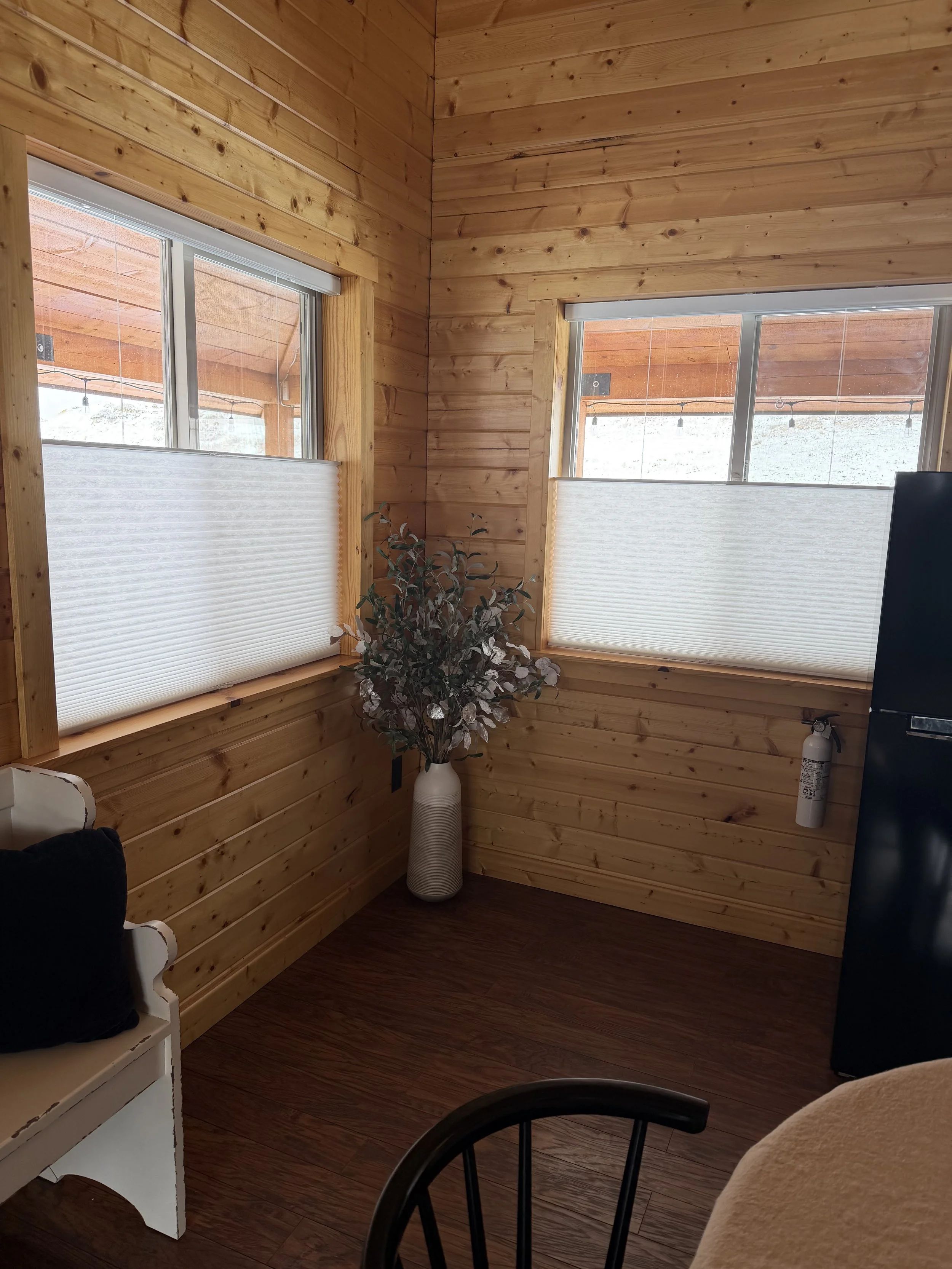 Corner of a wooden room with two windows covered with white blinds, a potted plant in a tall ceramic vase, part of a white bench with a black pillow, a black refrigerator, and a fire extinguisher mounted on the wall.