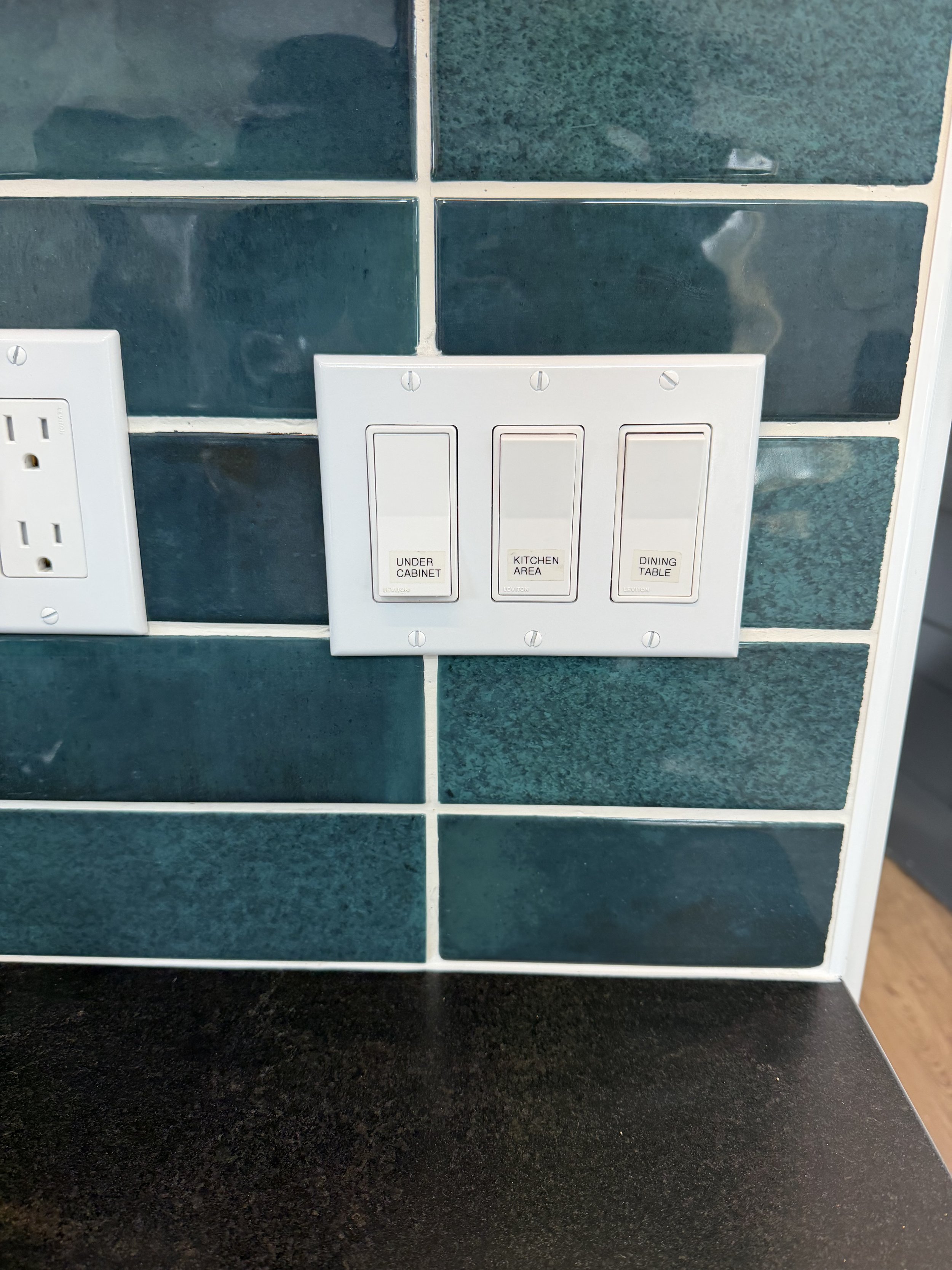 A white multi-switch panel with three labeled switches for under cabinet, kitchen area, and dining table, mounted on a blue-green tiled wall near electrical outlets.