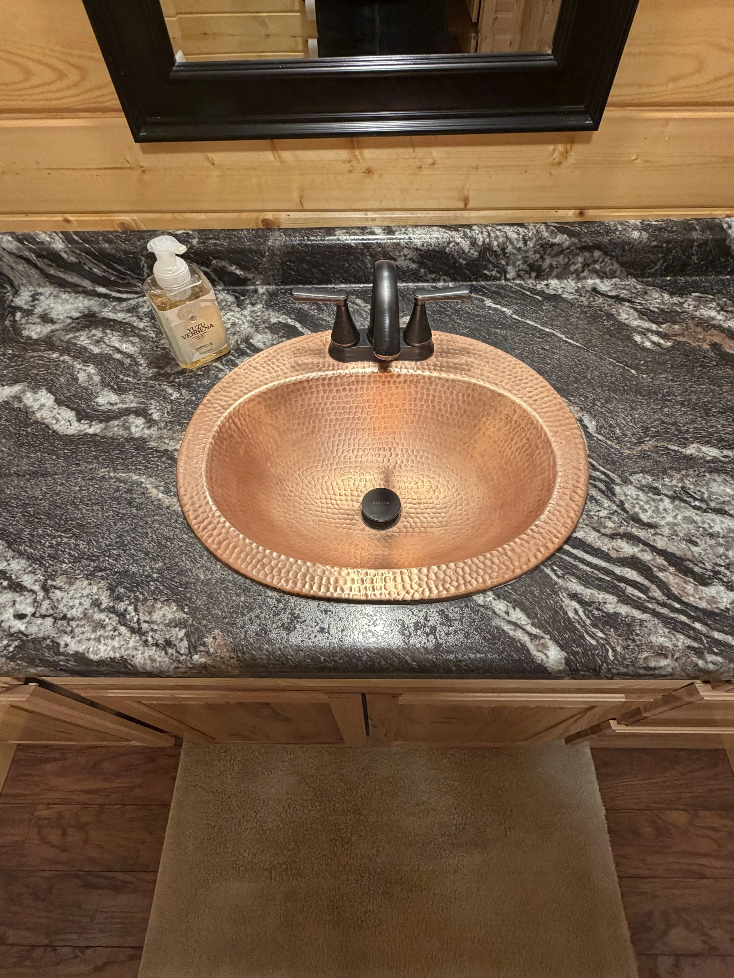 Copper sink with hammered texture, black faucet, soap dispenser on a dark granite countertop, mirror above, wooden cabinet below.