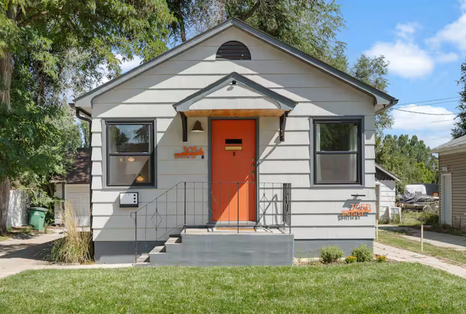 Front view of a small white house with an orange door, black metal railings, and two windows. Green lawn in the foreground, trees on sides, blue sky with some clouds in the background.