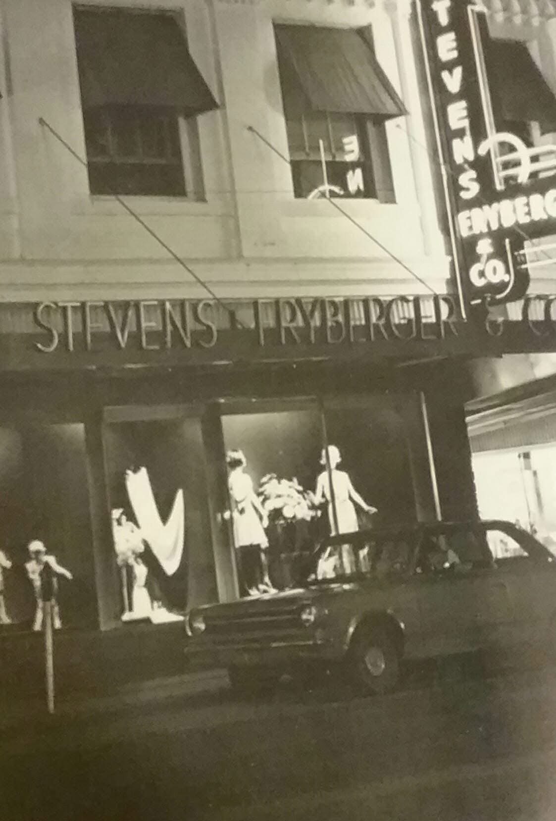 Black and white photo of a storefront with mannequins dressed in clothing, a car parked in front, and signs reading 'Stevens Pryberger' and 'Vans'.
