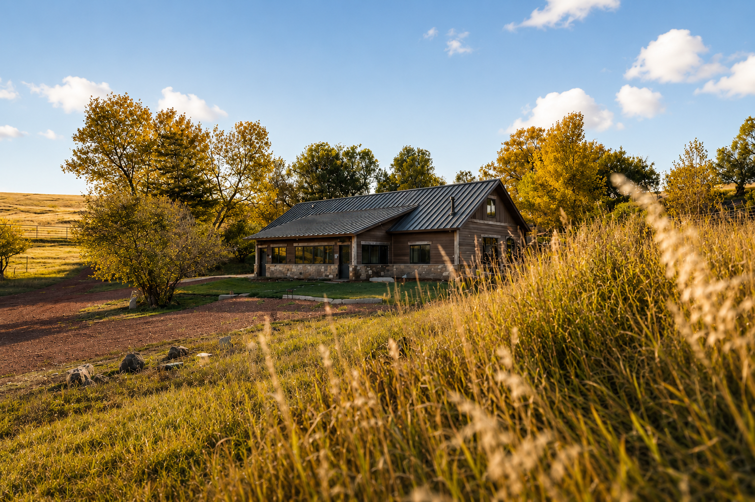 A rustic house with a metal roof situated on a grassy hill surrounded by trees and open landscape during golden hour.