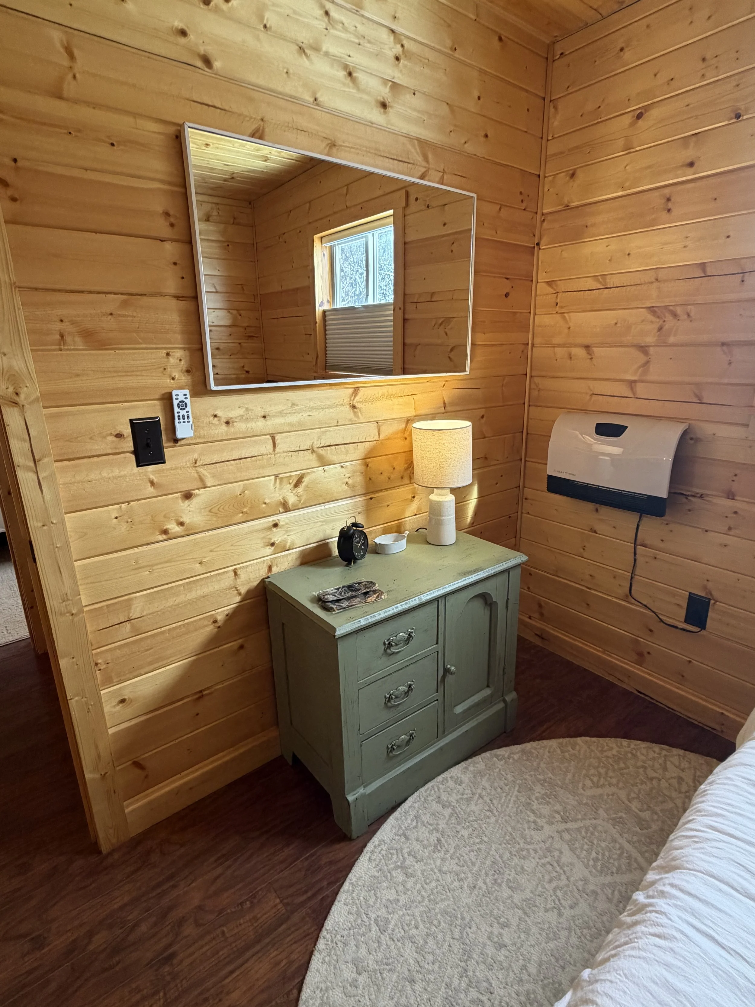 Interior of a cozy cabin bedroom with wooden walls, a small green nightstand, a mirror on the wall, a table lamp, an alarm clock, and a window with a view of trees.