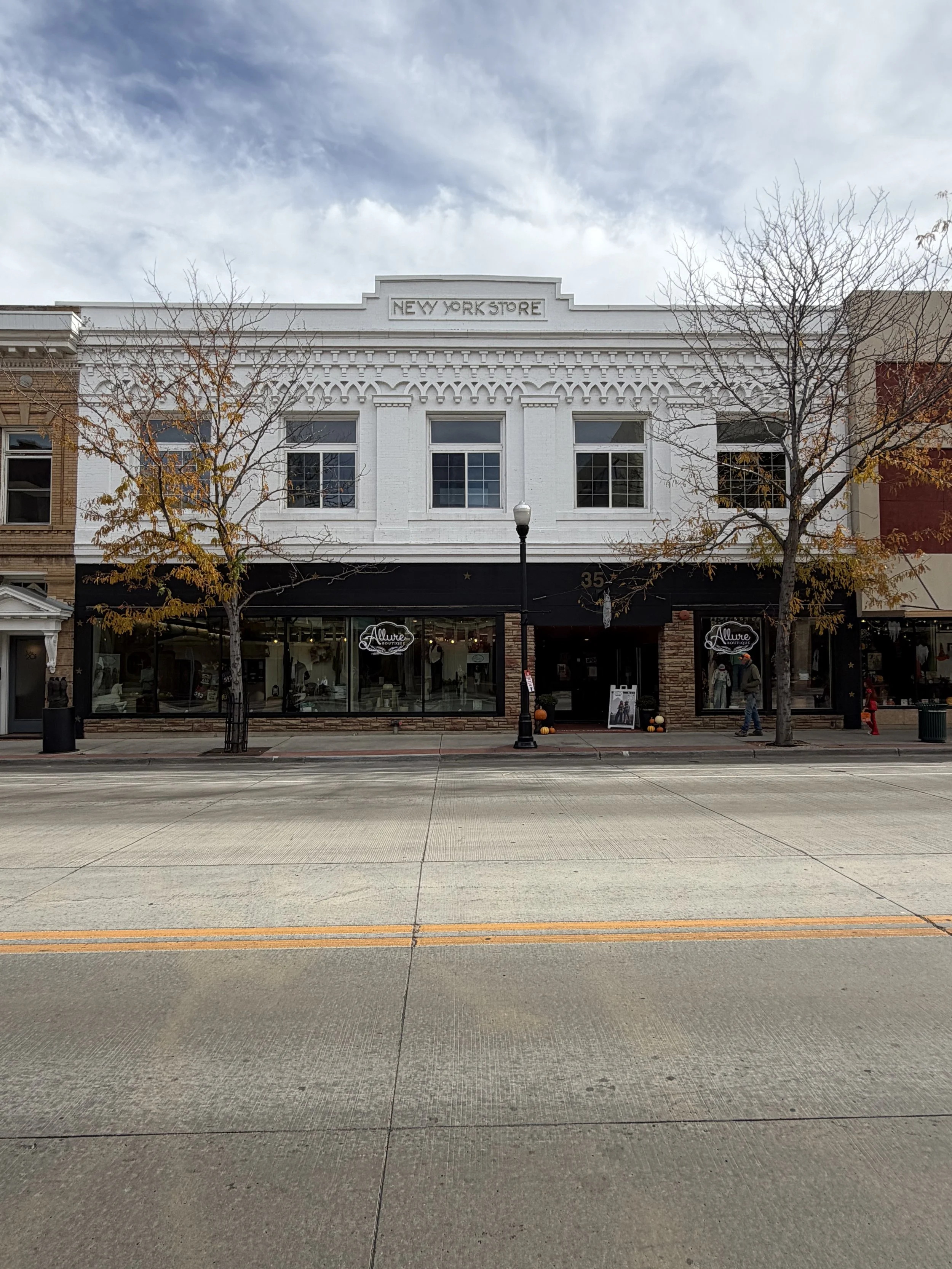 City street view with a white two-story building featuring decorative architectural details and a sign reading 'New York Store'; storefront at ground level with shoe store 'Allure Boutique'; leafless trees and pedestrians on the sidewalk; cloudy sky in the background.