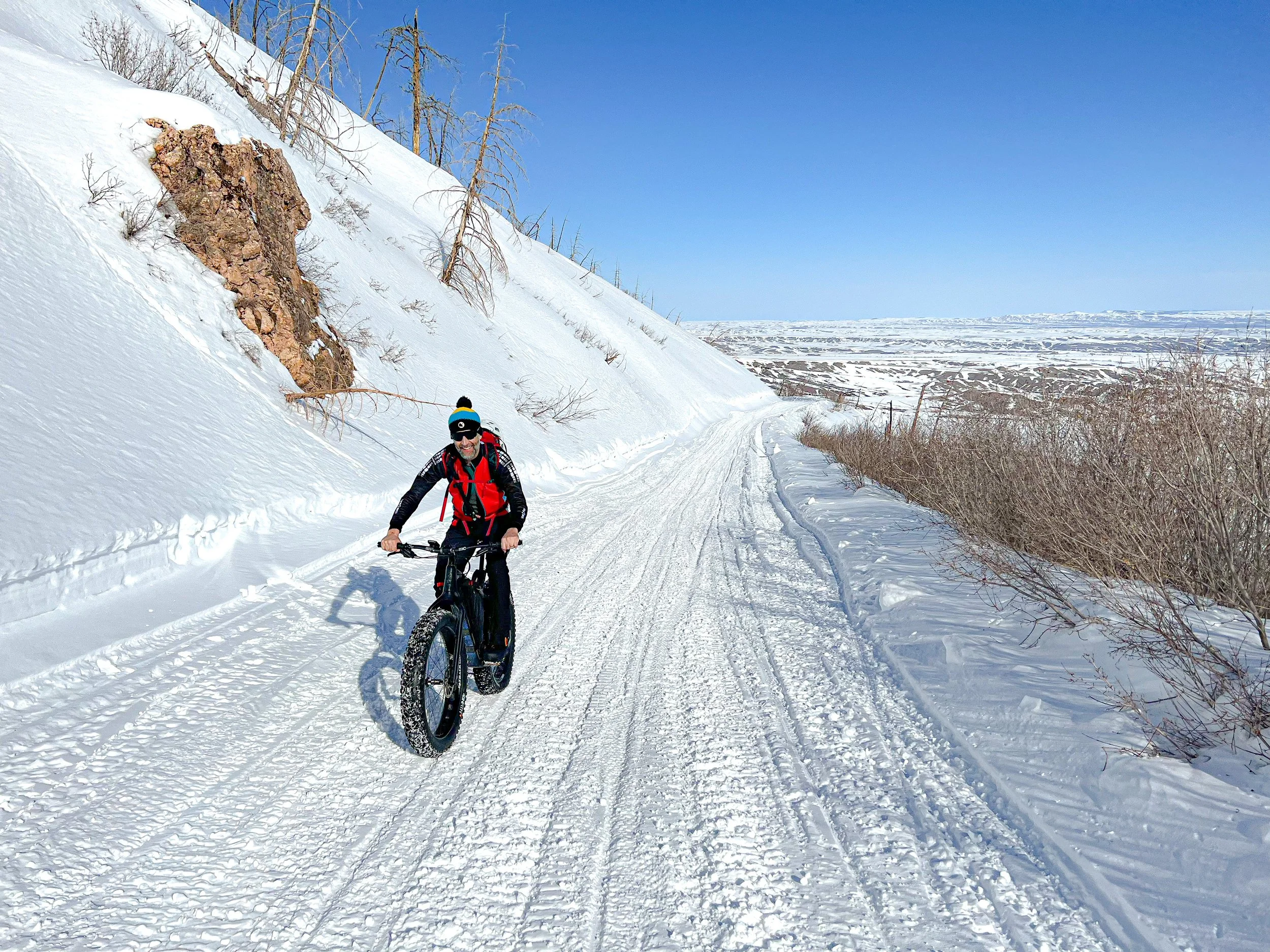 A man riding a fat tire bike on a snow-covered trail in a winter landscape with a clear blue sky.