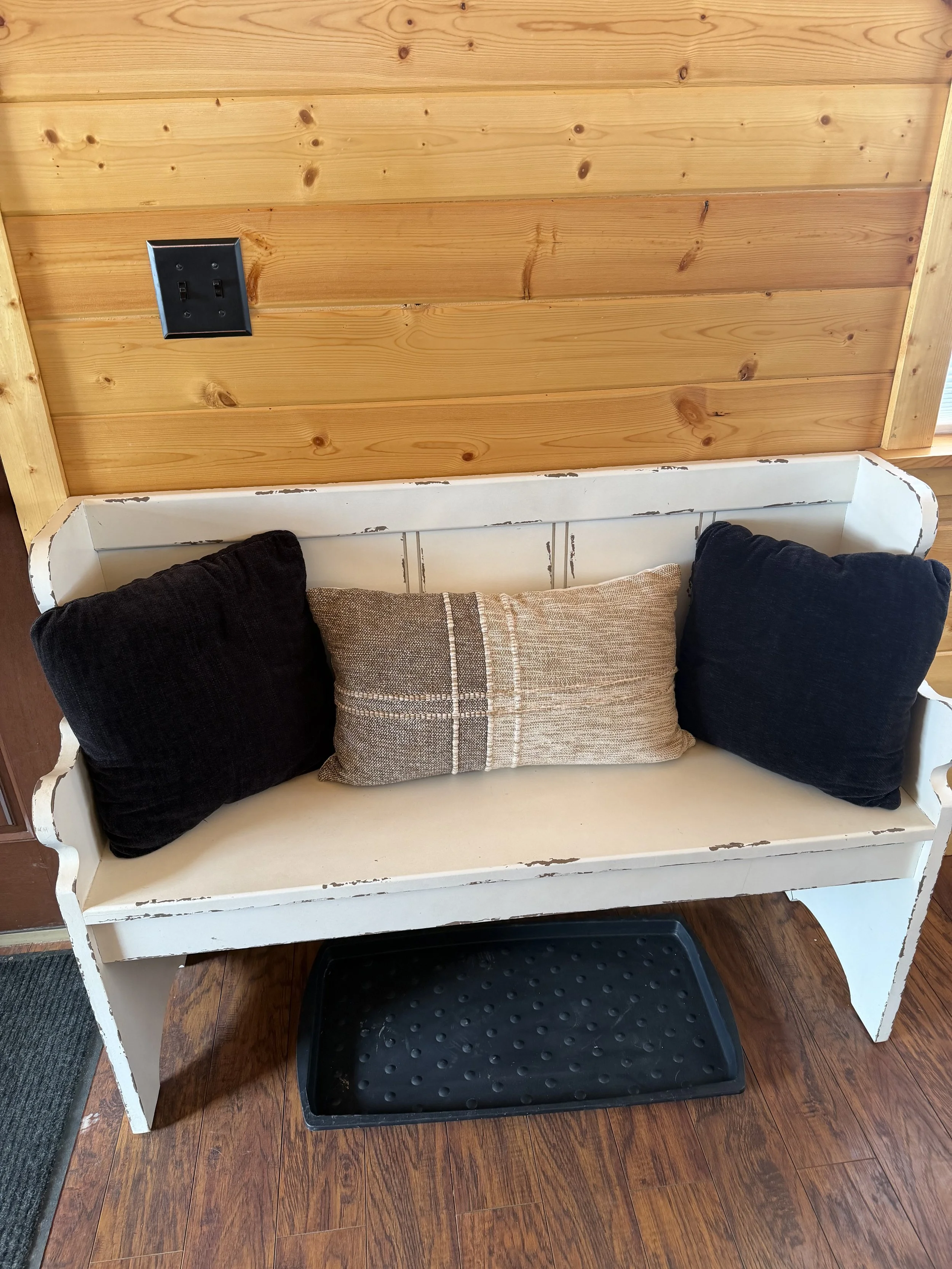 A white wooden bench with a distressed finish, featuring ornate armrests, positioned against a wooden-paneled wall, with a black rubber tray underneath and two black pillows and one rectangular striped pillow on the bench.