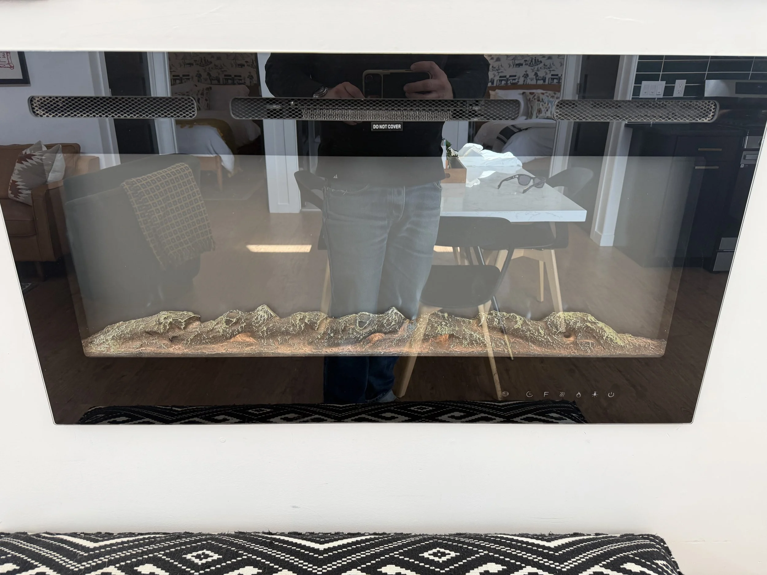 A modern fireplace with a glass front and a landscape design inside, reflecting a person taking a photo in a room with furniture and decor.