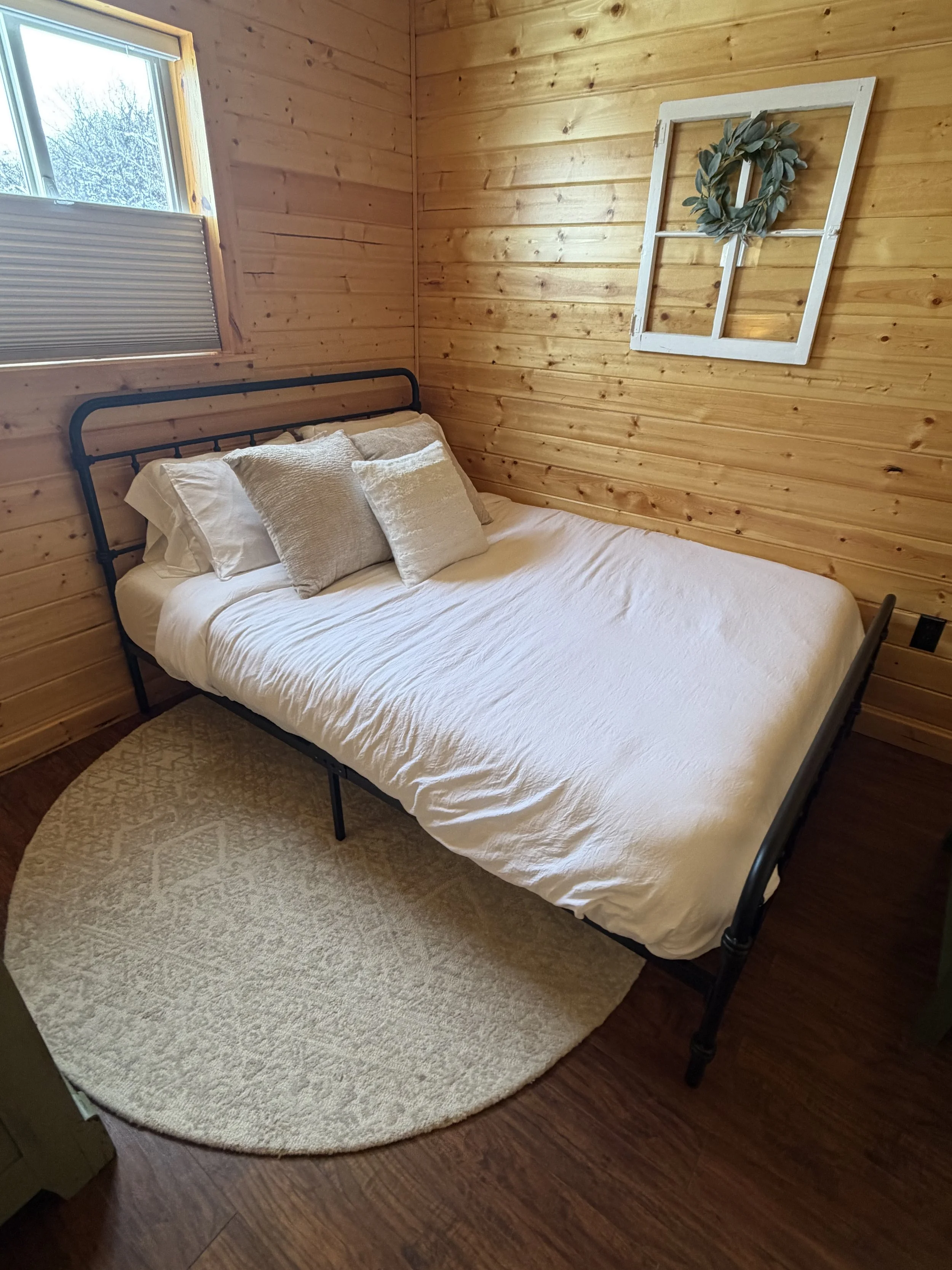 A cozy bedroom with wood paneled walls, a black metal bed frame, white bedding, and three decorative pillows. A round beige rug is placed under the bed, and a small window with a view of trees is on the left side.
