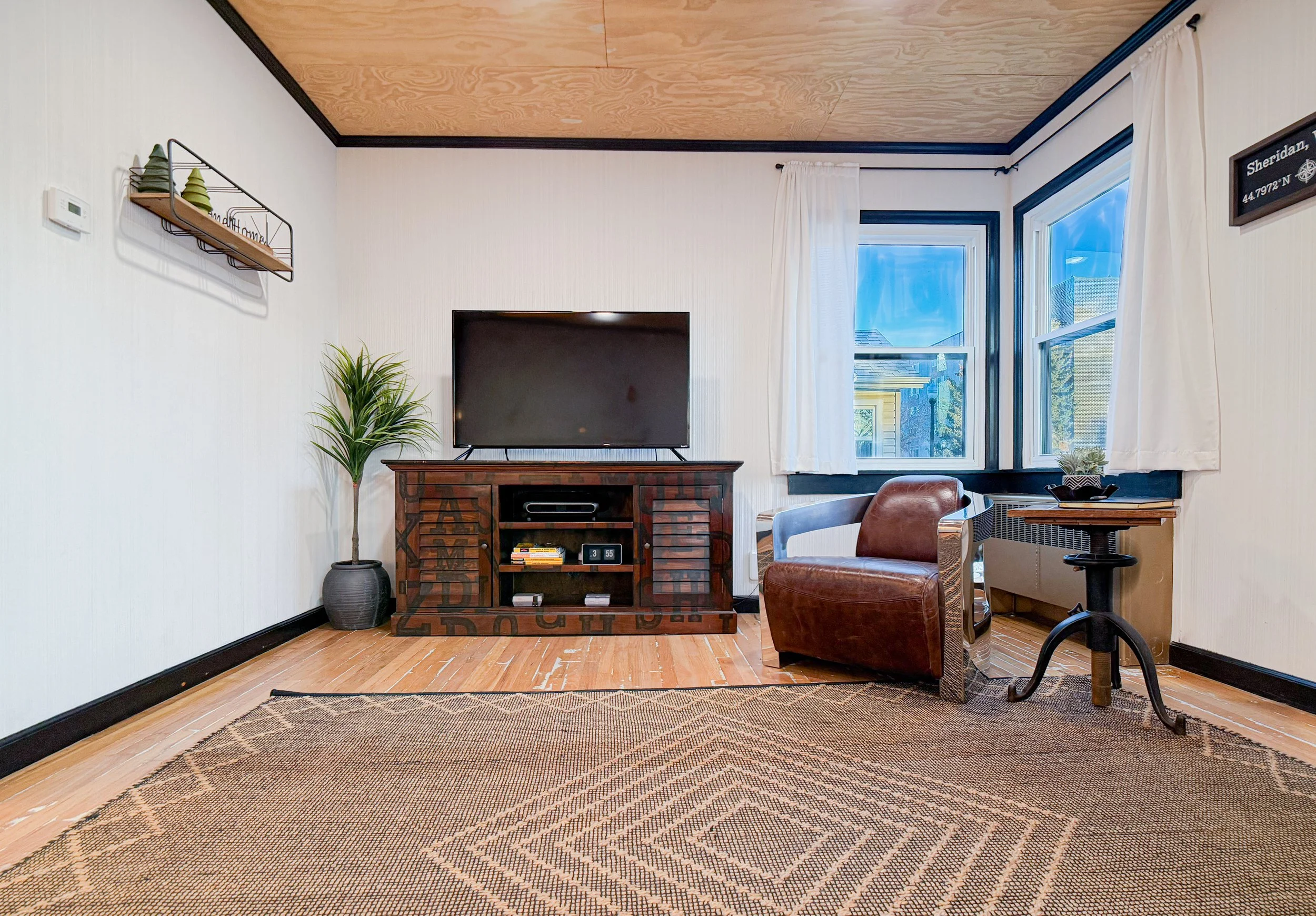 Living room with a large flat-screen TV on a wooden stand, a potted plant to the left of the TV, a leather armchair, a small round side table with a potted plant, a patterned area rug, and a window with white curtains showing a view of neighboring houses and trees.