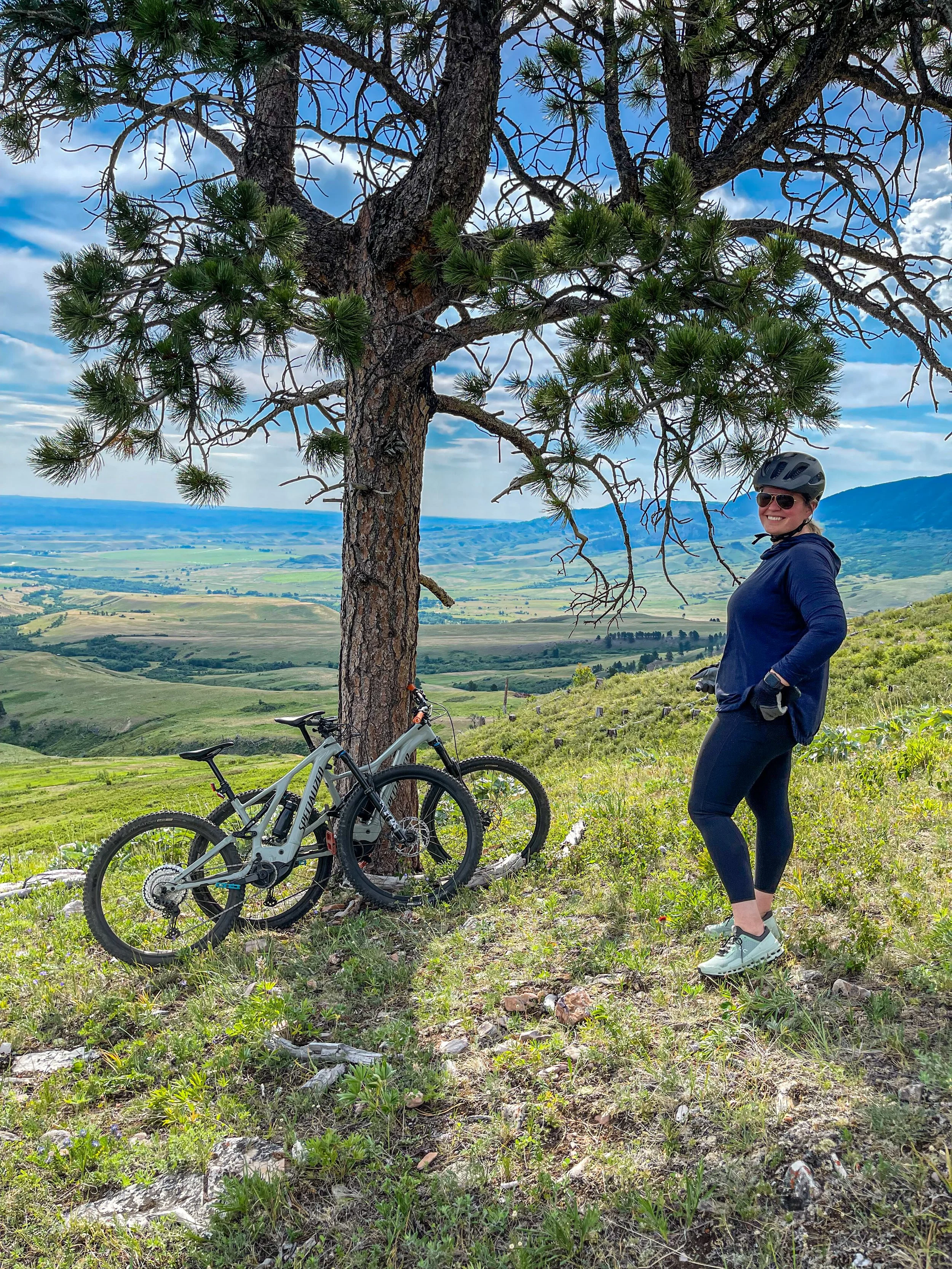 A woman in athletic clothing and a helmet standing with hands on hips next to two mountain bikes leaning against a tree on a hilltop, overlooking a lush green valley with a blue sky and scattered clouds.
