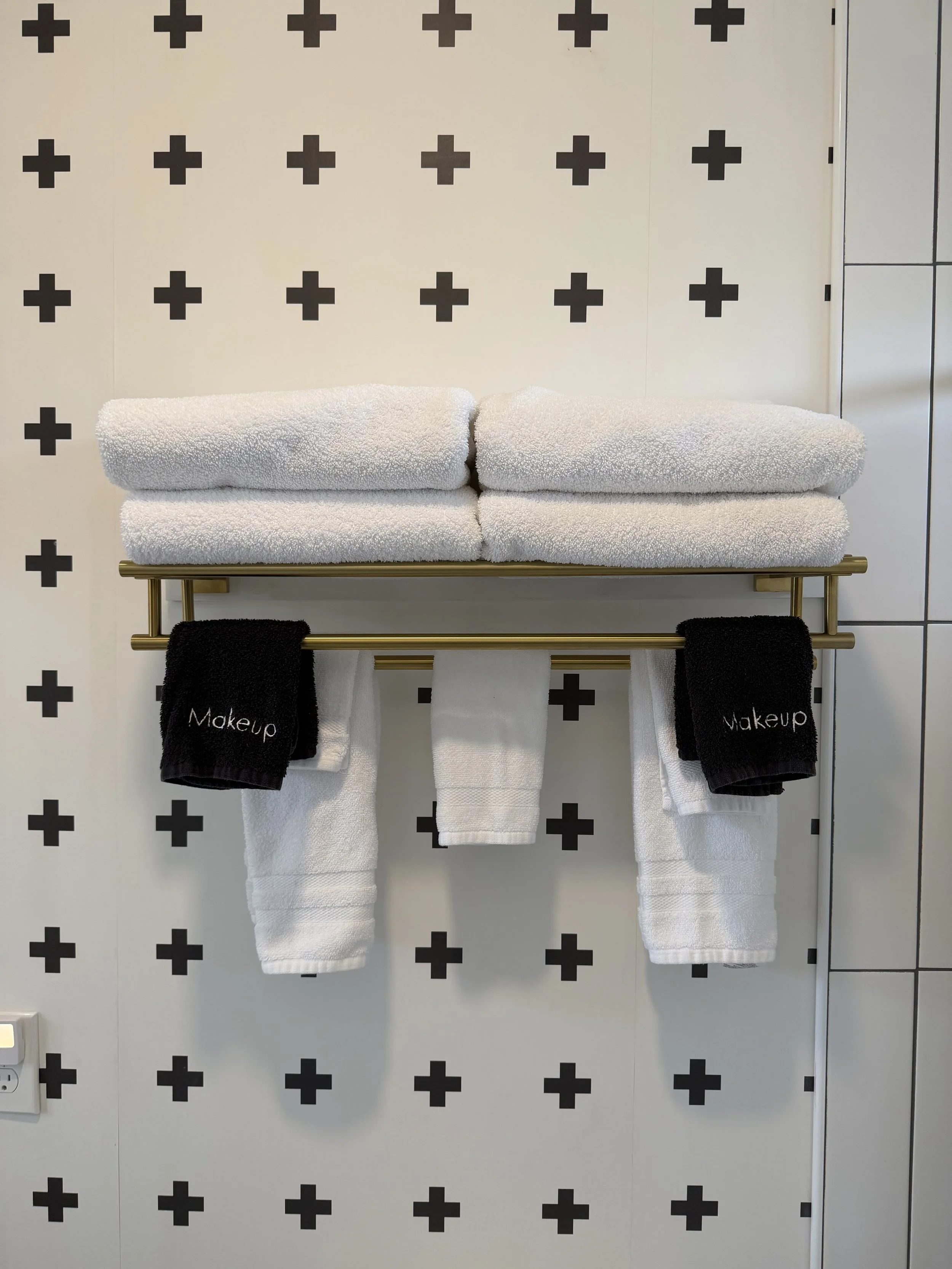 Stacked white towels on a shelf and hanging towels labeled 'Makeup' in a bathroom with black cross-patterned wallpaper.