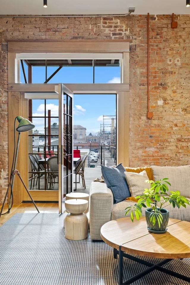 Living room with a beige couch, blue and beige pillows, a round wooden coffee table with a green potted plant, a standing lamp, and glass double doors opening to a balcony with outdoor chairs, a table, and a view of buildings and a bright blue sky.
