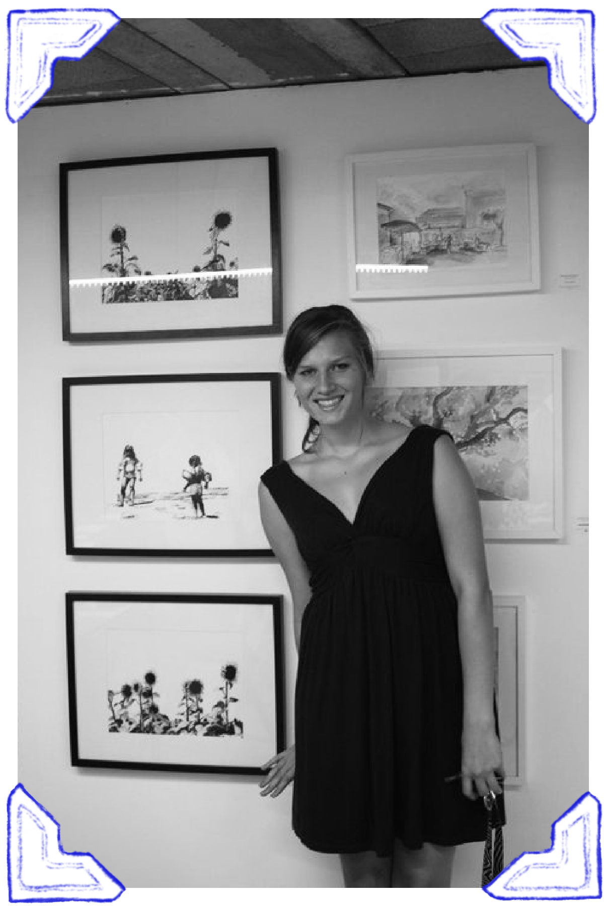 A woman in a black dress standing in front of a white wall with framed artwork, smiling at the camera.