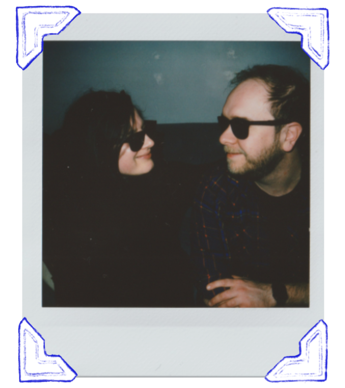 A woman and a man wearing sunglasses are looking at each other and smiling in a dimly lit space.