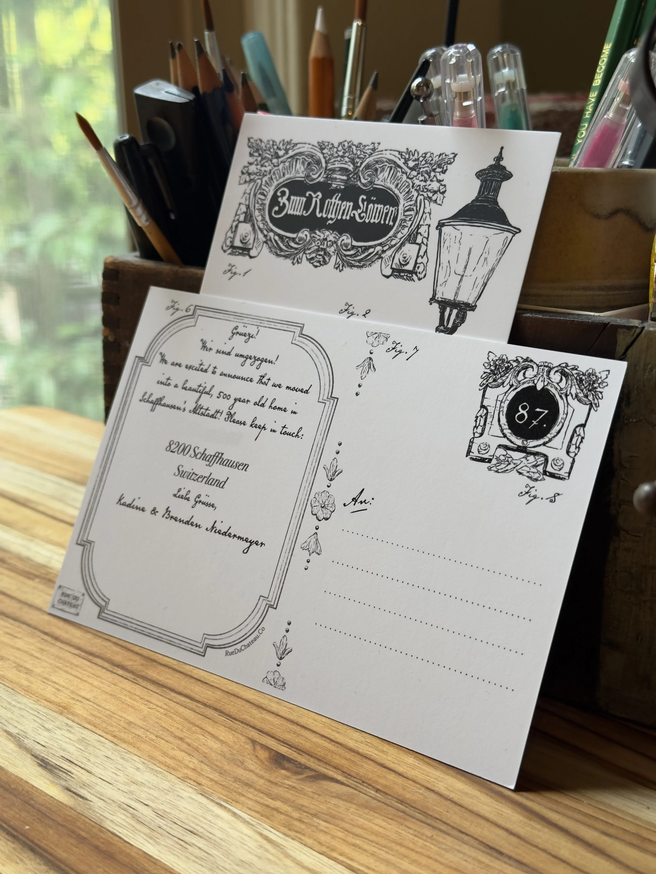 A white paper with decorative English words and illustrations of street lamps and floral borders on a wooden surface, with a container of pens and pencils in the background.