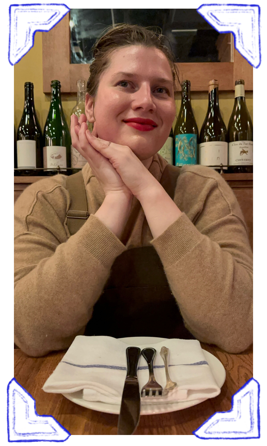 A woman with short hair and red lipstick sitting at a restaurant table, resting her chin on her hands, with wine bottles in the background.