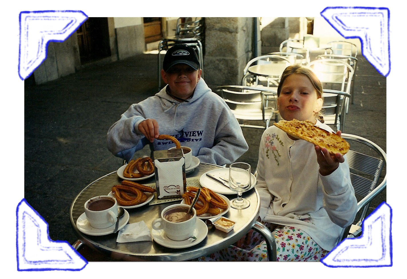 Two children sitting at an outdoor cafe table enjoying breakfast with coffee, churros, and a large slice of pizza, with empty chairs in the background.