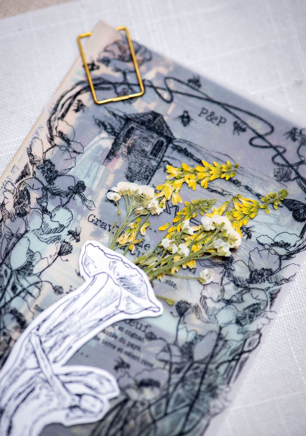 A floral arrangement of yellow and white flowers placed on a sheet of paper with black line drawings of flowers and a building, held down by a yellow paperclip, with a black and white sticker of a flower at the bottom.