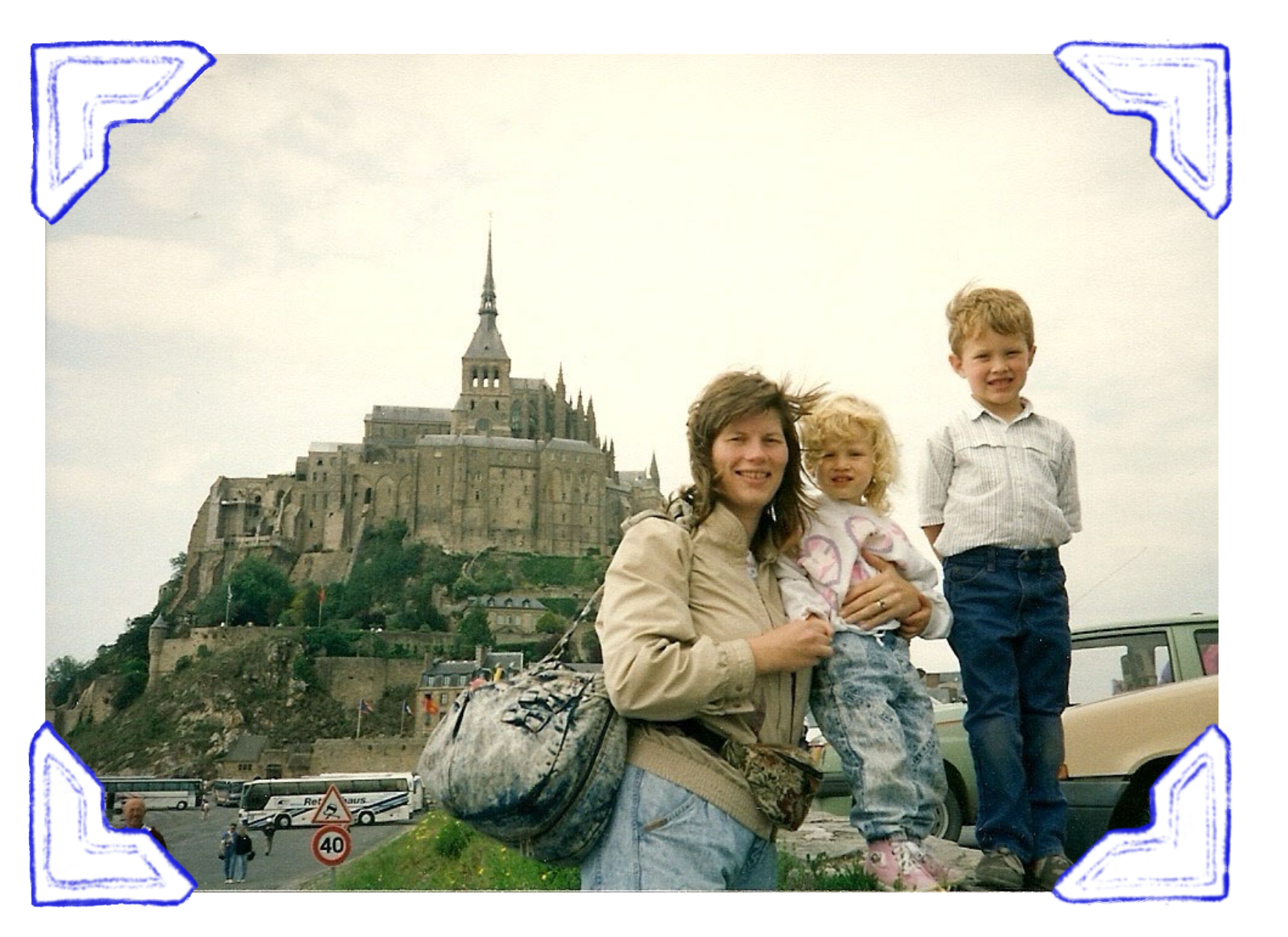 A woman with two young children standing in front of Mont-Saint-Michel, a historic island commune in France, with a parking lot and traffic signs in the foreground.