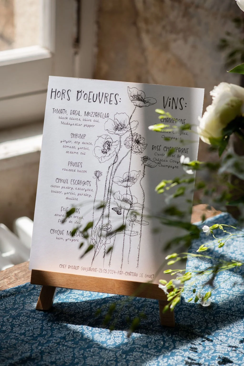 A handwritten menu on white paper with floral illustrations, listing hors d'oeuvres and wines, displayed on a wooden stand on a blue patterned cloth, with some white flowers in the foreground.