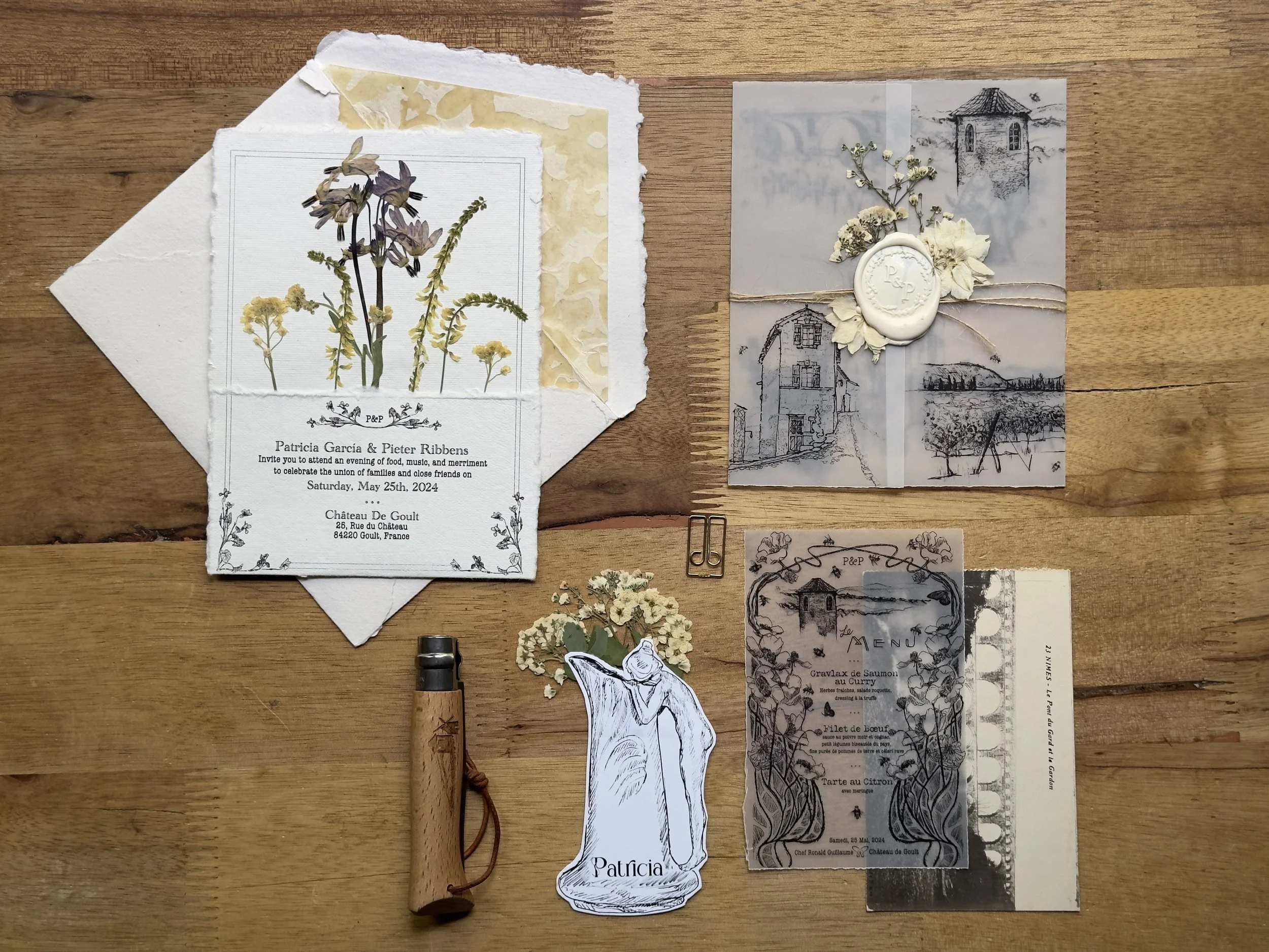 Flat-lay of wedding or event stationery including invitation cards, menus, and decorative dried flowers displayed on a wooden surface. There's also a branding stamp, a small tool that looks like a corkscrew, and a paperclip.