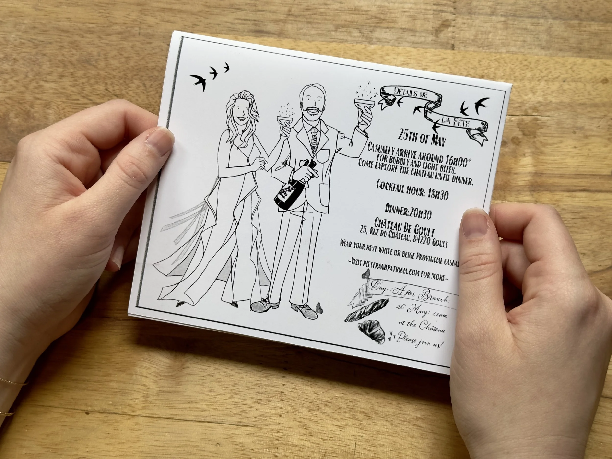 A hand holding a black-and-white illustrated flyer on a wooden table. The flyer features drawings of a woman and a man holding drinks, with flying birds in the background and decorative elements. The text on the flyer mentions details of a celebration event on May 25th, including arrival, dinner, and drinks, with a location in Goult, France.