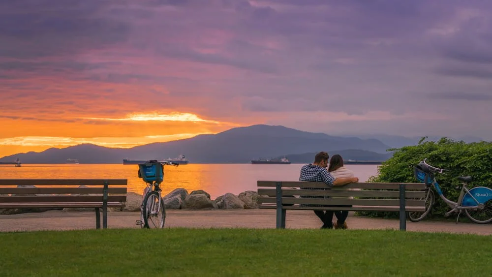 A couple on a park bench by the water at sunset in Vancouver, with mountains behind