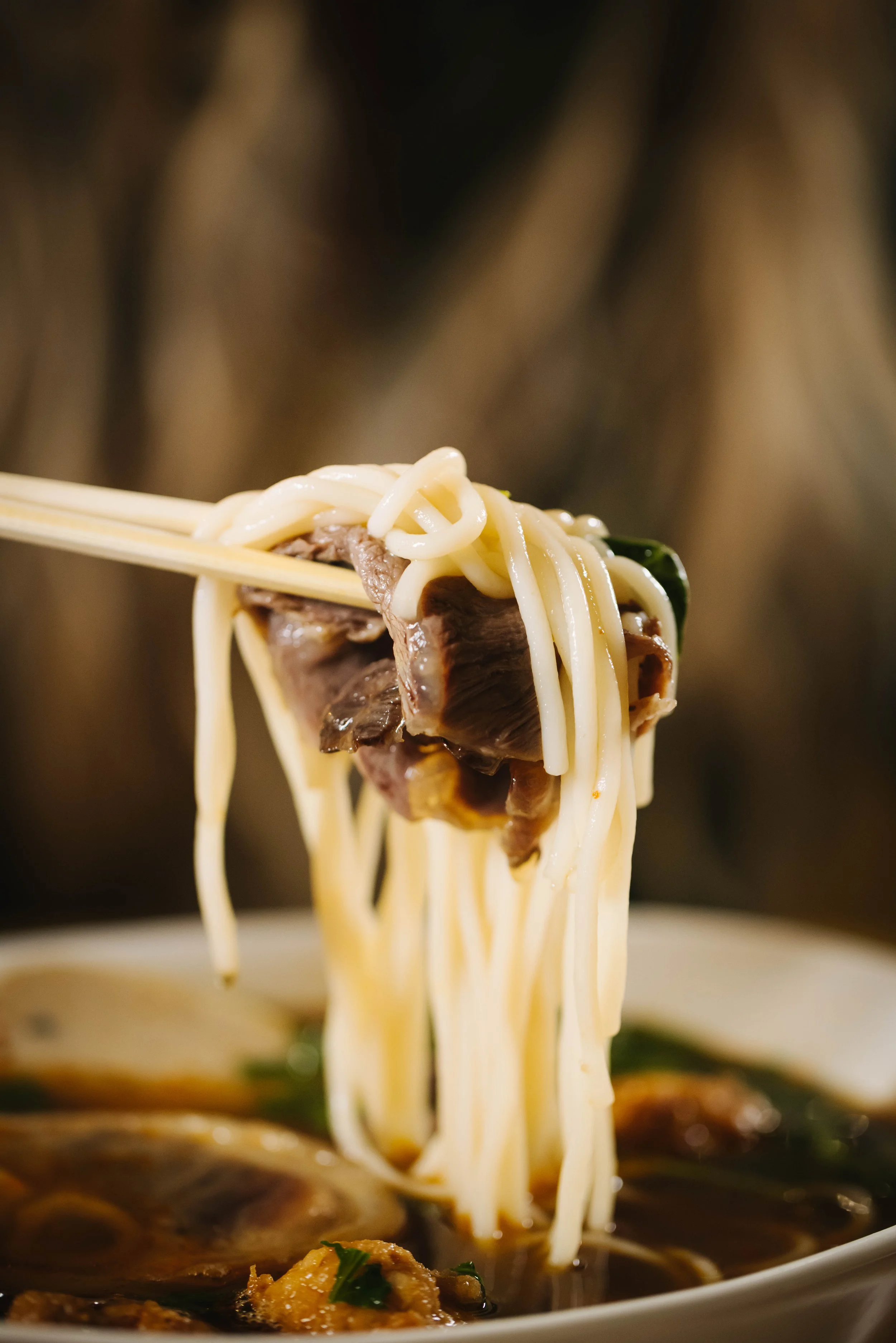 Close-up of chopsticks holding noodles, beef, and vegetables in a bowl of ramen.