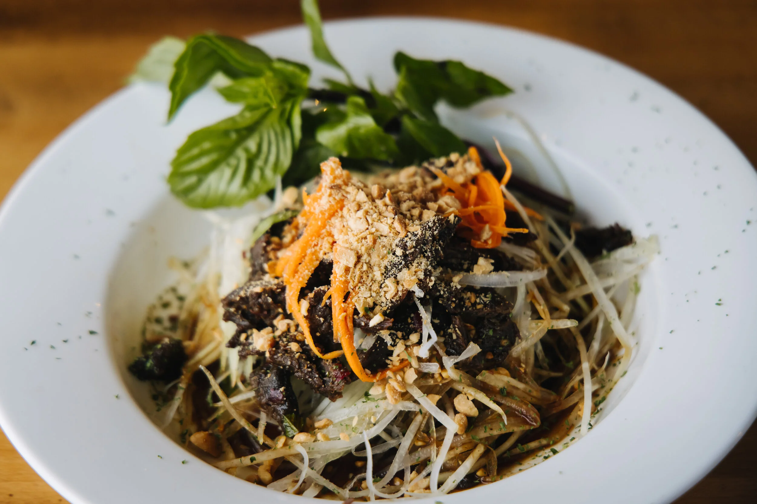 A white bowl filled with a Vietnamese-style noodle salad topped with shredded carrots, crushed peanuts, and fresh herbs.