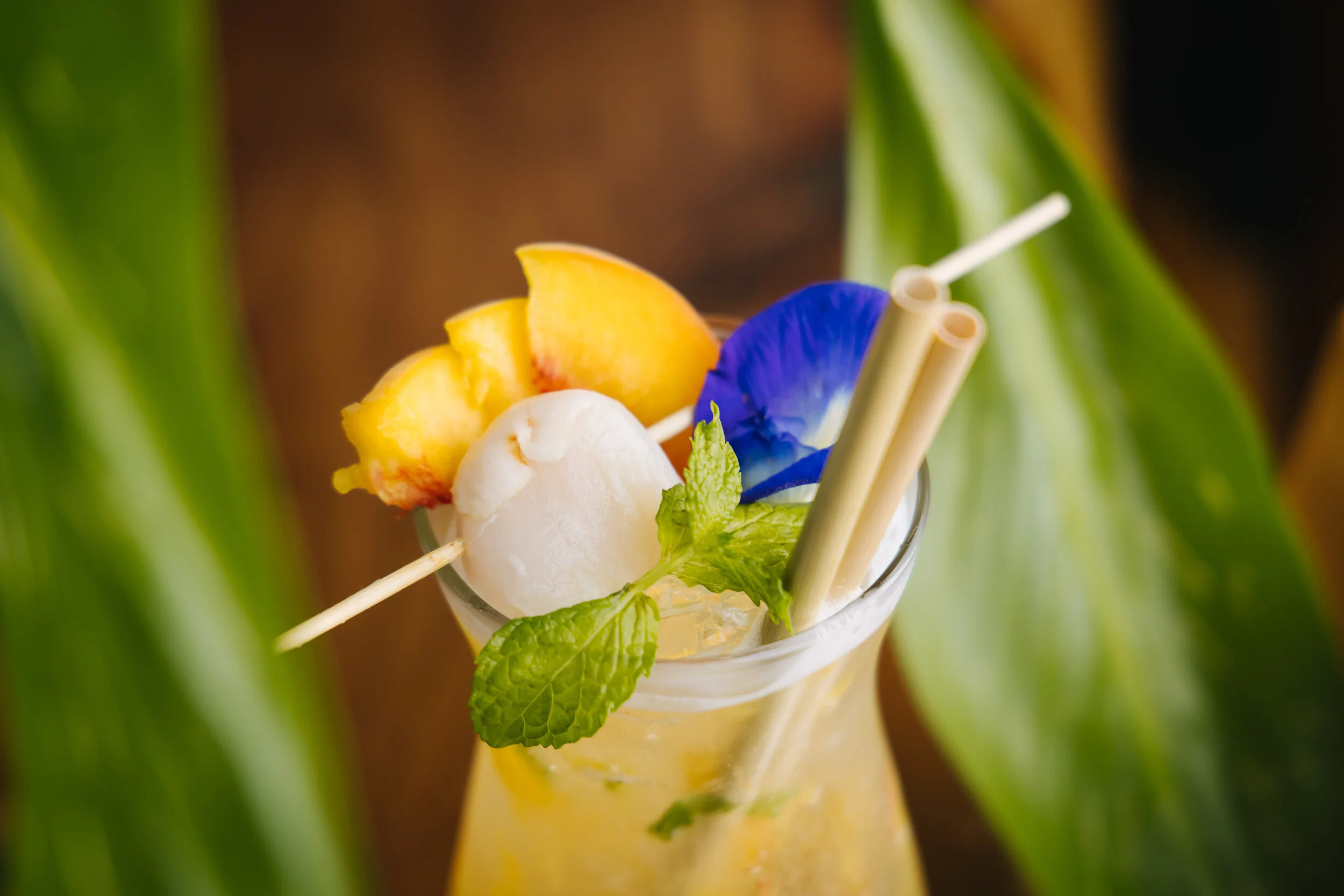 A glass of tropical fruit cocktail garnished with a slice of peach, a scoop of ice cream or frozen fruit, a blue edible flower, and fresh mint leaves, with straw-like stirrers inside.