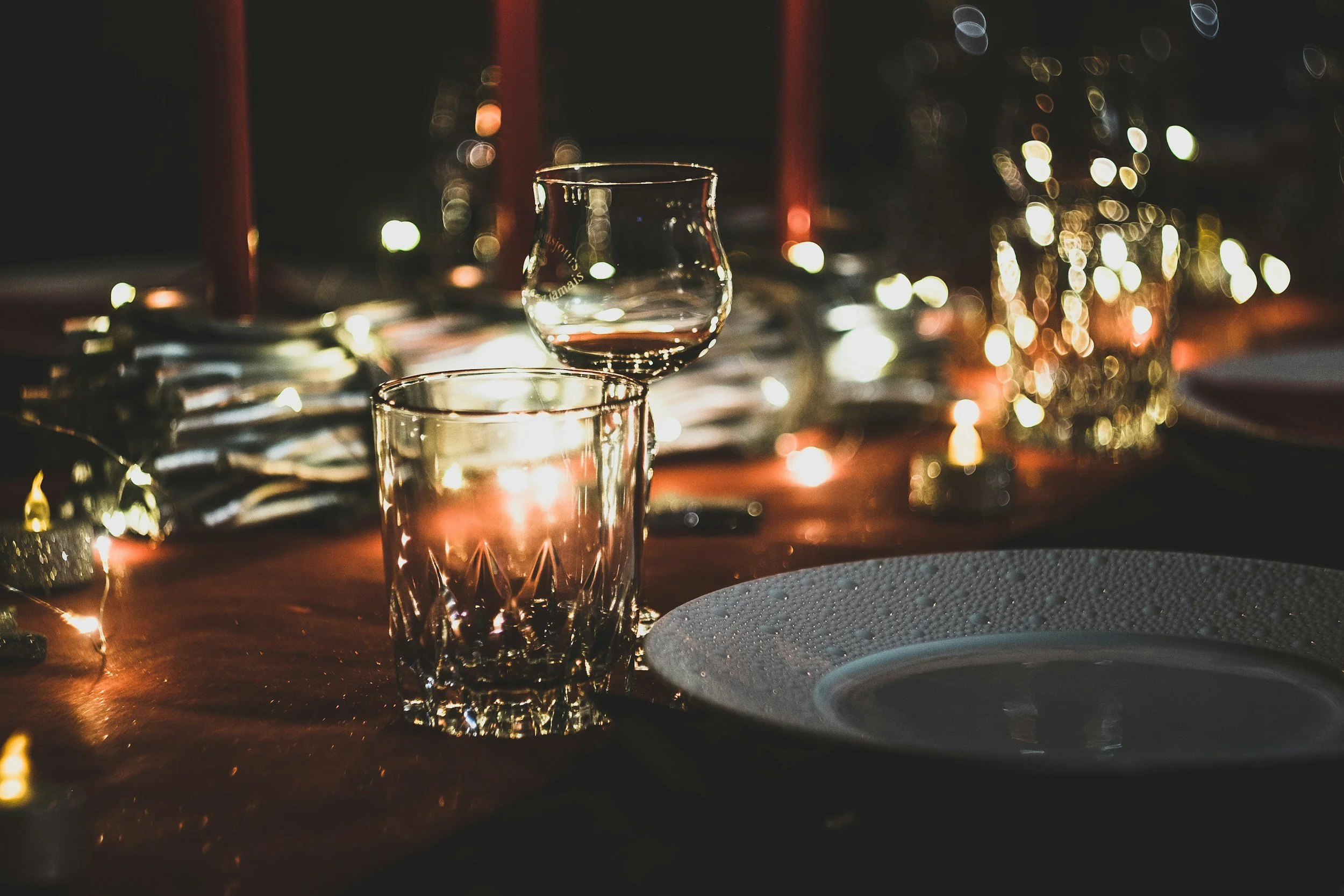 A table decorated for a celebration with a lit candle, a short glass with a drink, and a snifter glass with a darker drink, with blurred warm lights in the background.