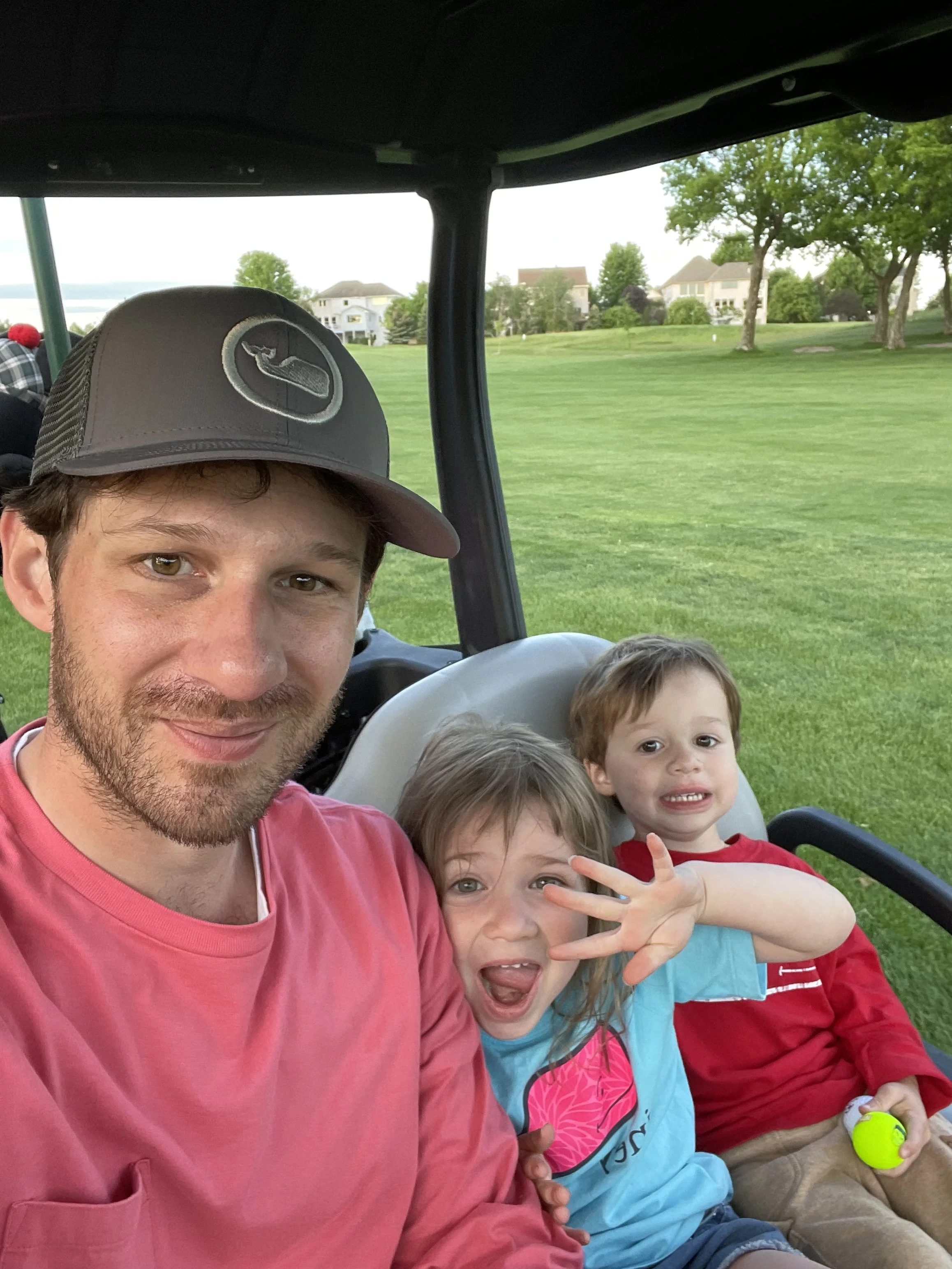 A man and two children are sitting in a golf cart on a golf course with houses and trees in the background.