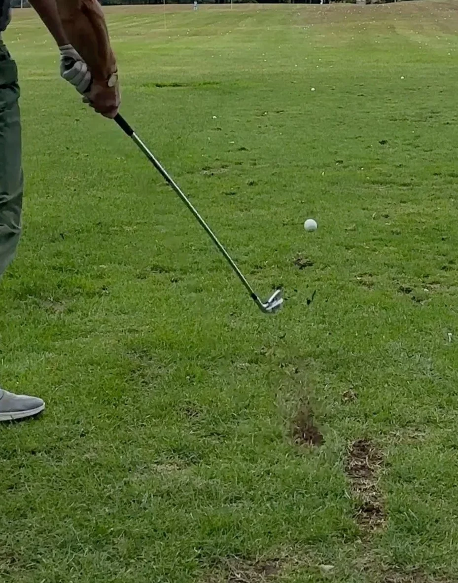Golfer hitting a ball at a driving range just after impact, with divots visible in the turf.
