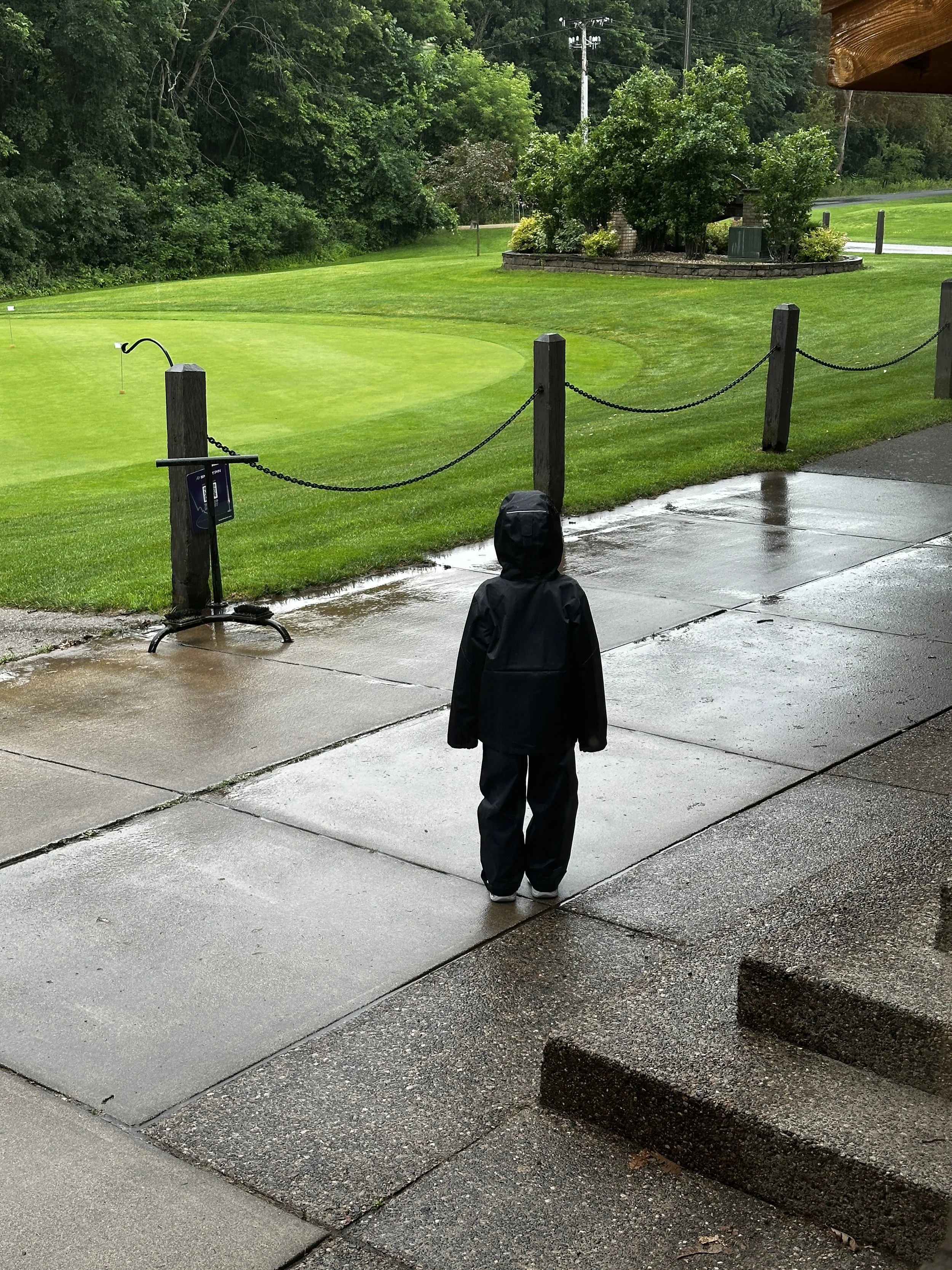 A young child dressed in a black raincoat and rain pants standing on a wet sidewalk, looking at a golf course on a rainy day.