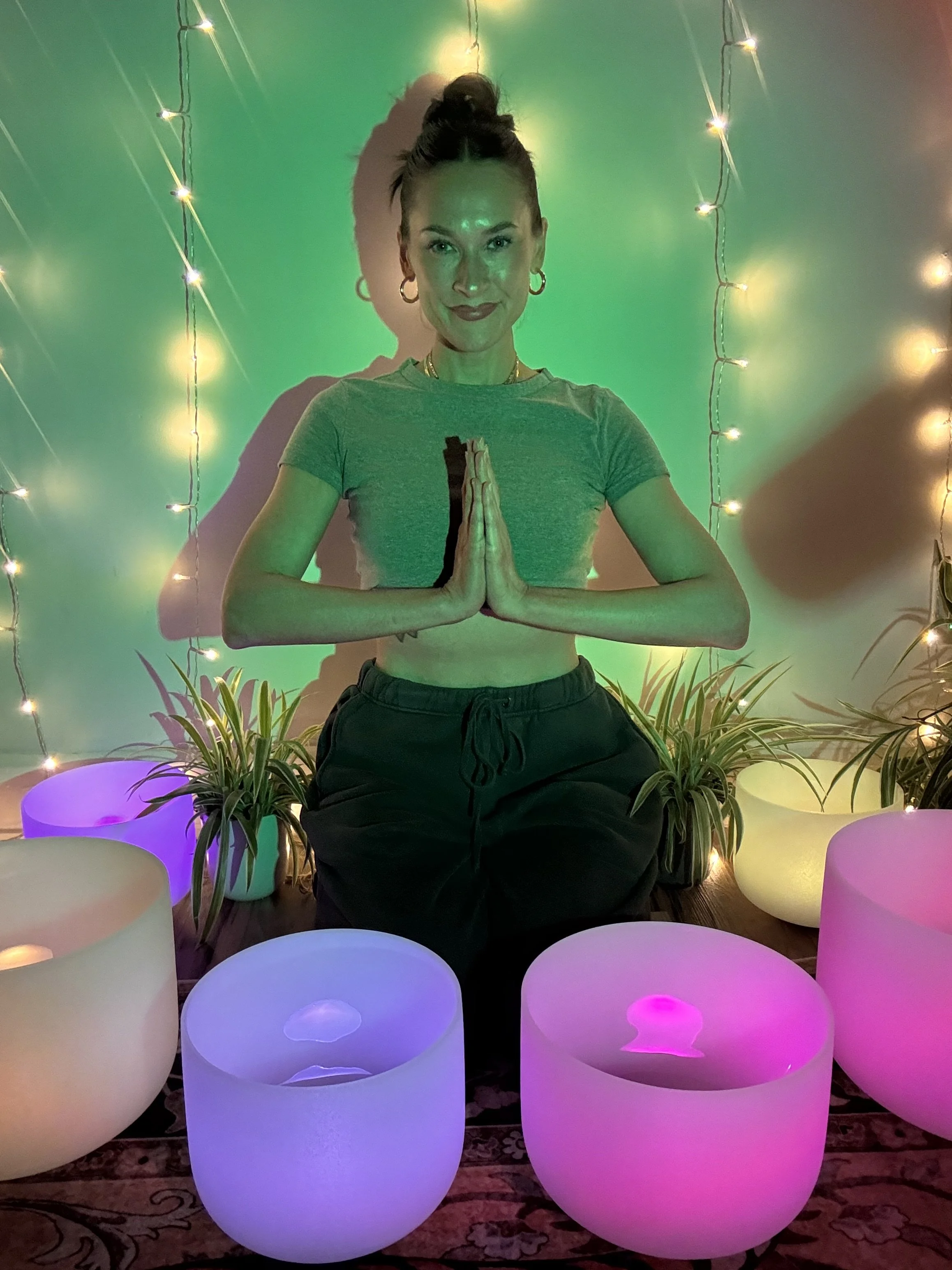 A woman practicing yoga with hands pressed together in front of her chest, surrounded by colorful illuminated lights, plants, and glowing lanterns in a decorated space.