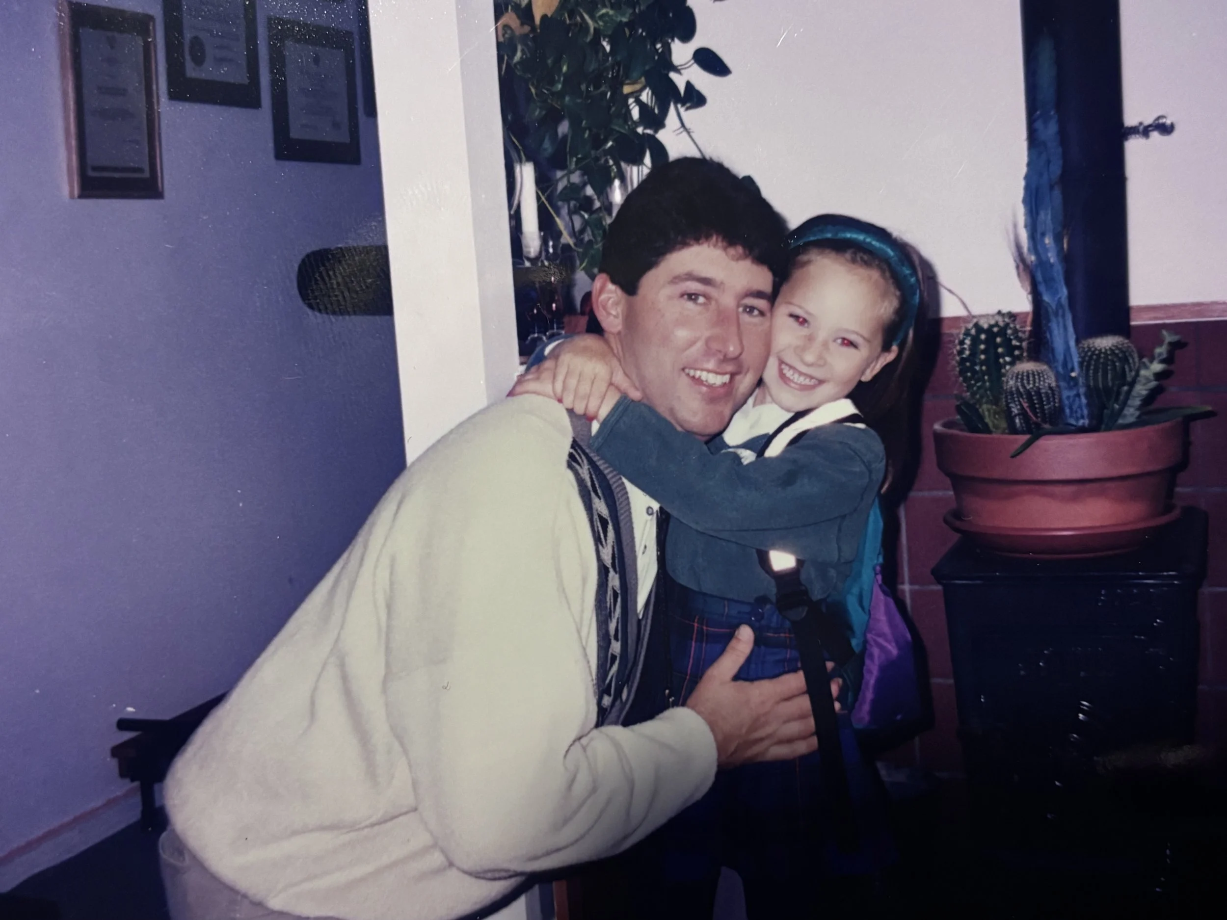 A man and a young girl embracing and smiling at the camera in an indoor setting. The man is seated, wearing a beige jacket, and the girl is wearing a blue headband and a dark jacket. There are potted cacti and framed pictures on the wall in the background.