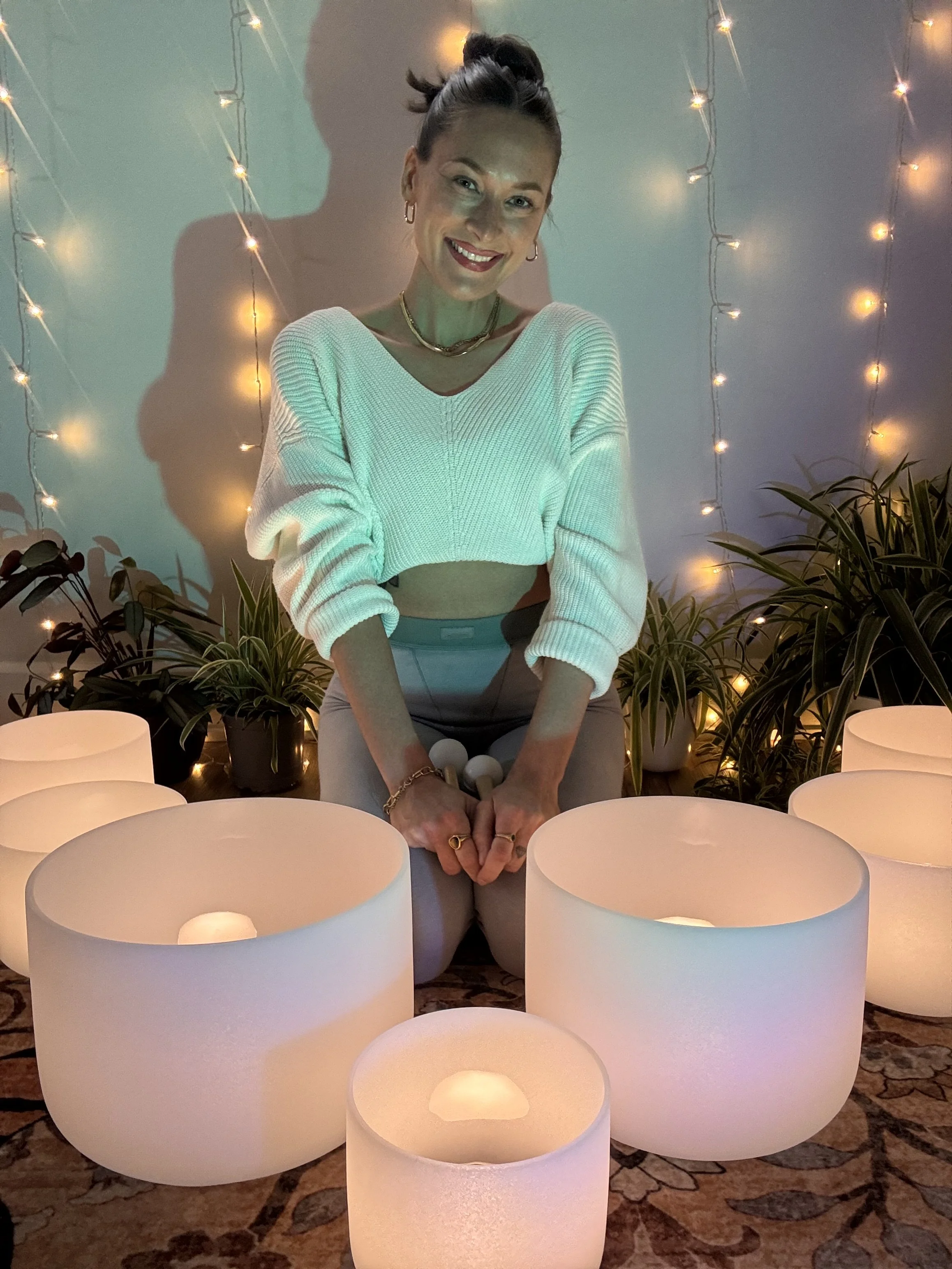 A woman with her hair in a bun, wearing a white sweater and multiple necklaces, smiling in front of a wall decorated with string lights and surrounded by white cylindrical candles and plants.