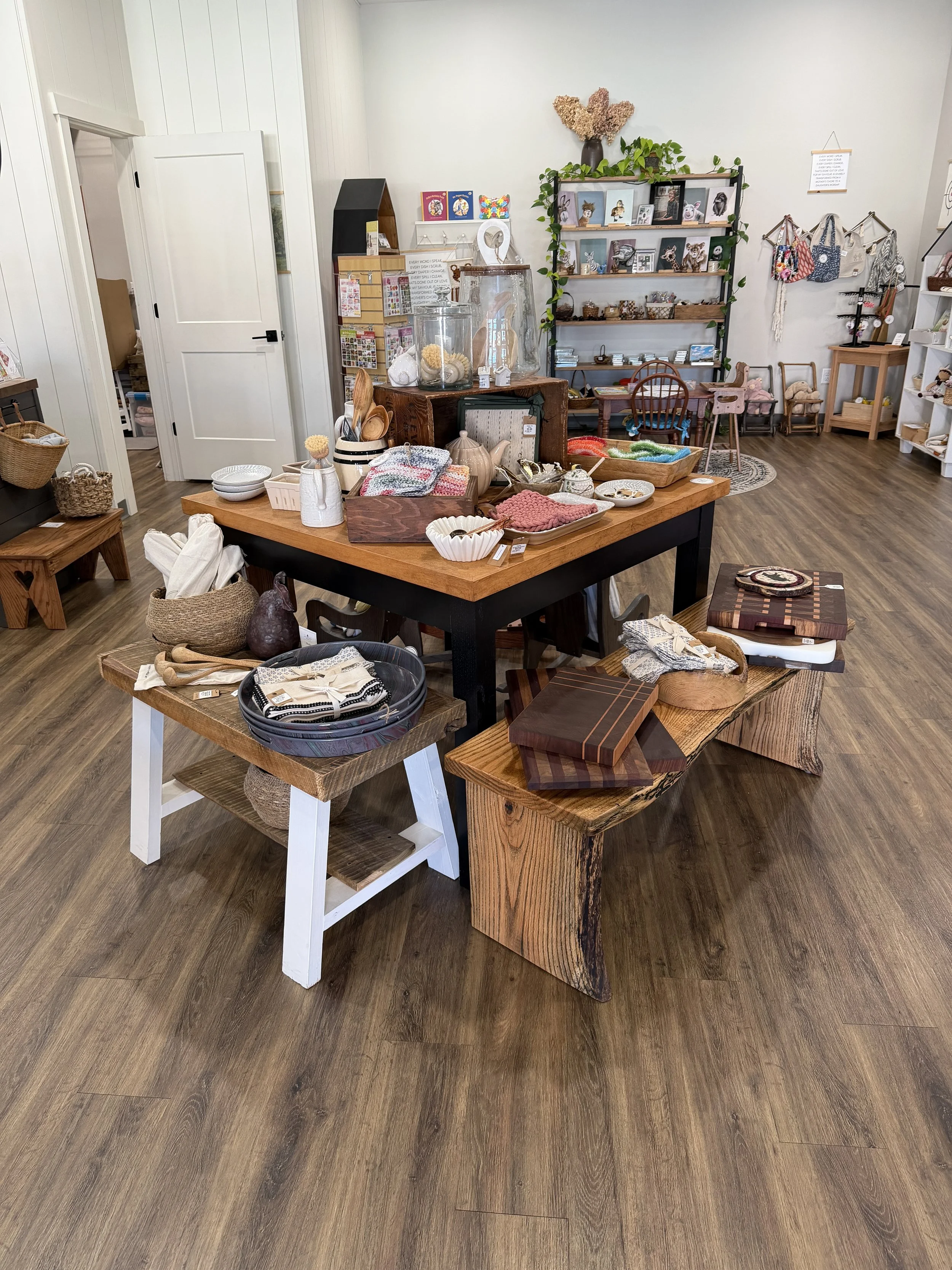 Interior of a retail store with wooden floors displaying various home decor and gift items on tables and shelves, including ceramics, textiles, picture frames, and decorative objects.