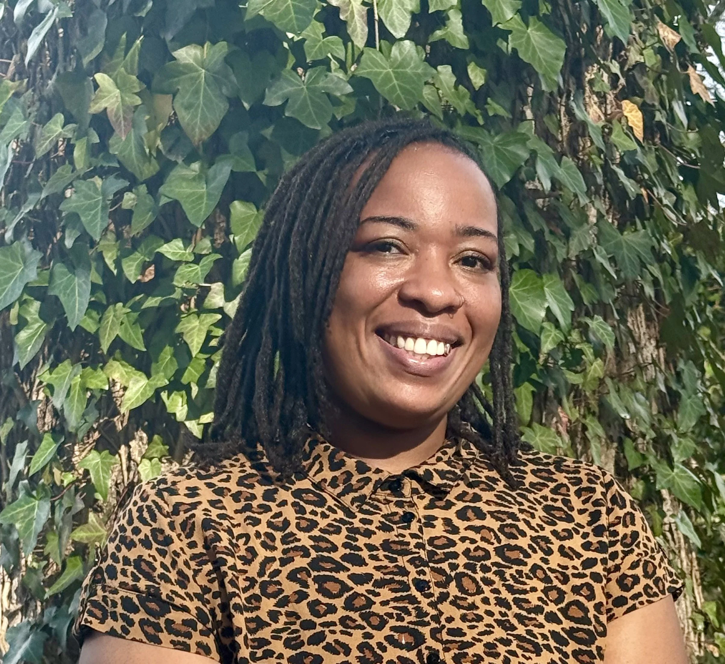 A woman with dark skin and long dreadlocks smiling in front of green ivy leaves, wearing a leopard print top.
