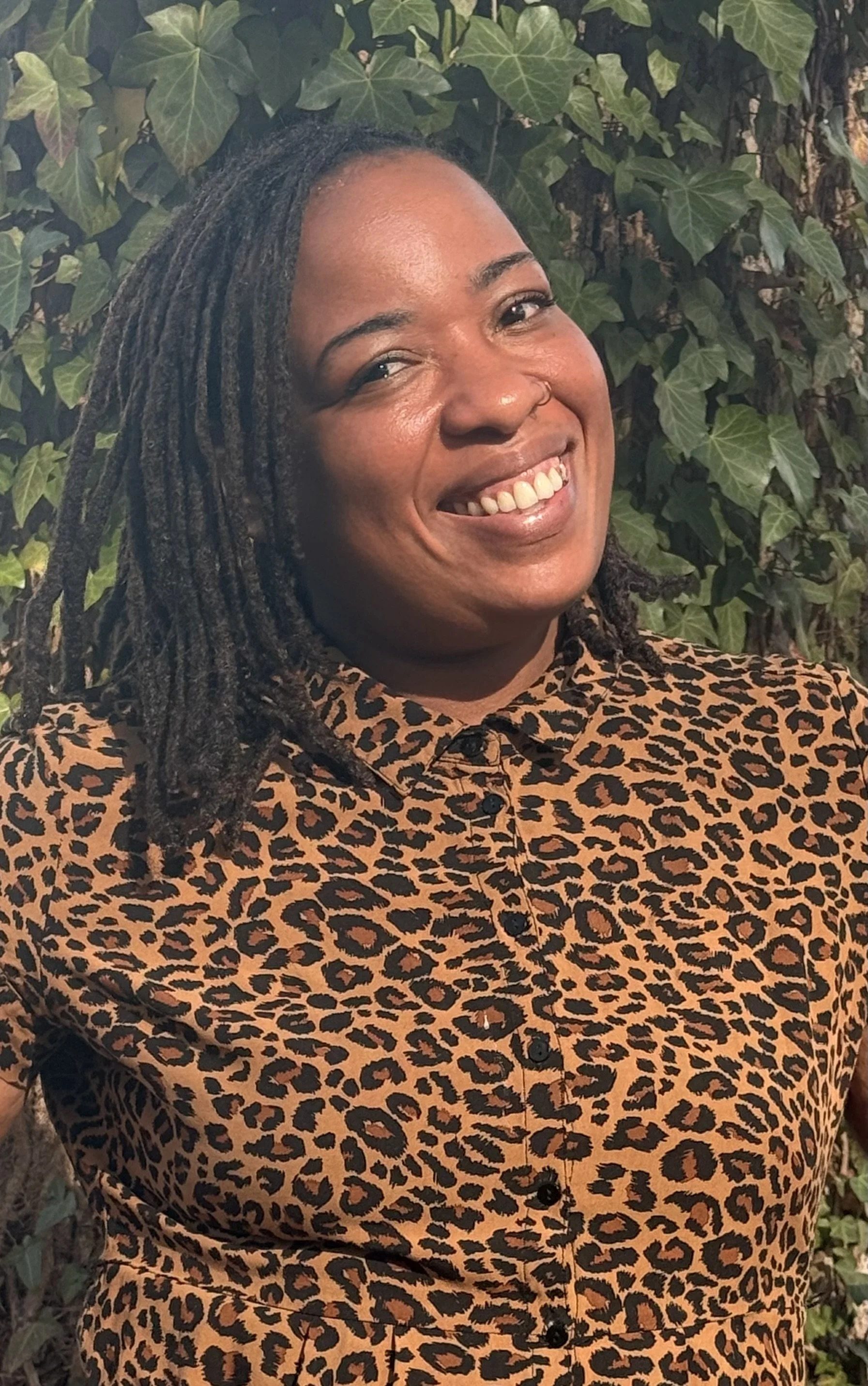 Close-up of a smiling woman with dreadlocks, wearing a leopard-print shirt, standing in front of green ivy leaves.