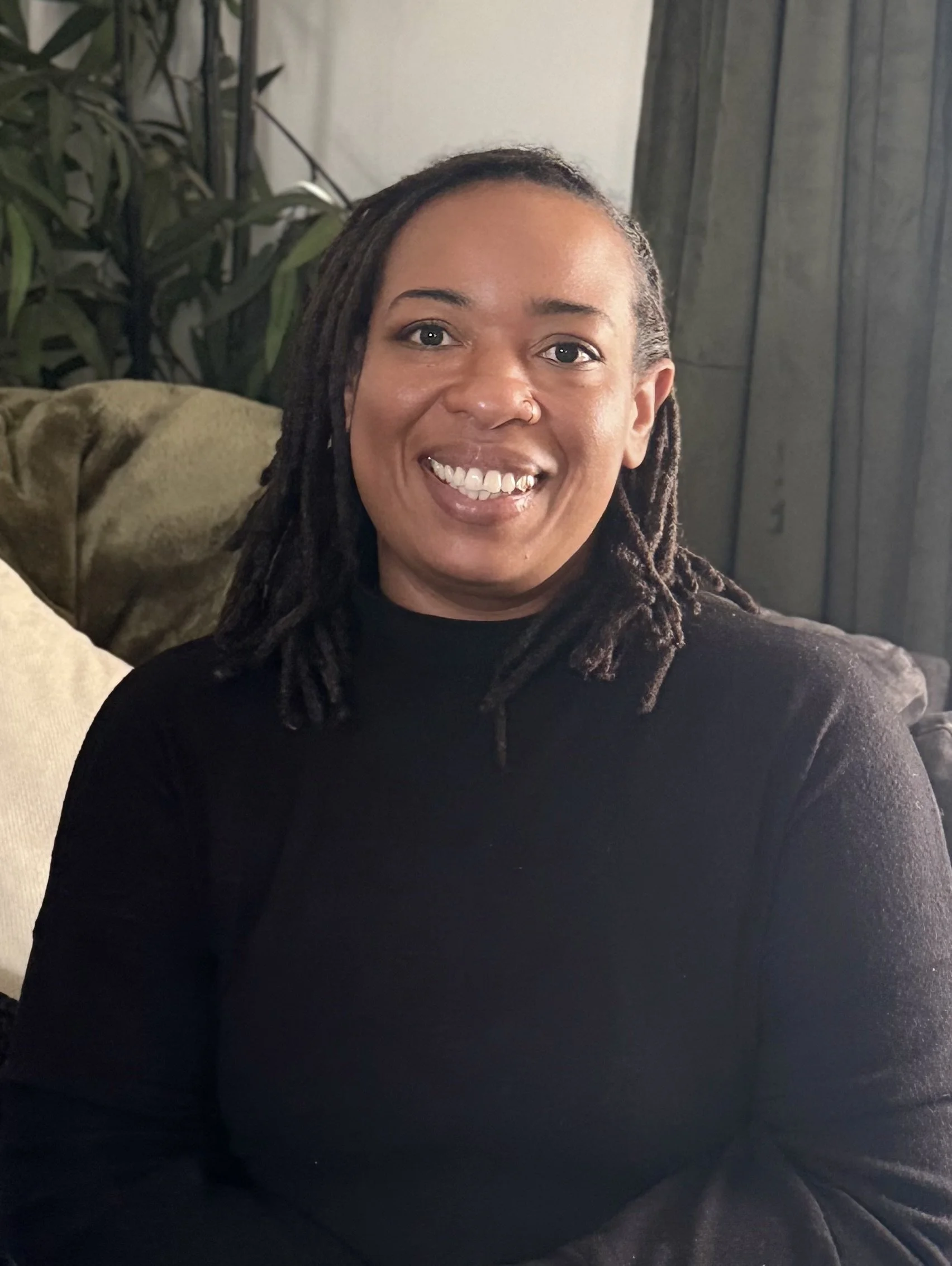 A smiling woman with dreadlocks sitting on a beige couch in a cozy living room with dark curtains and plants in the background.