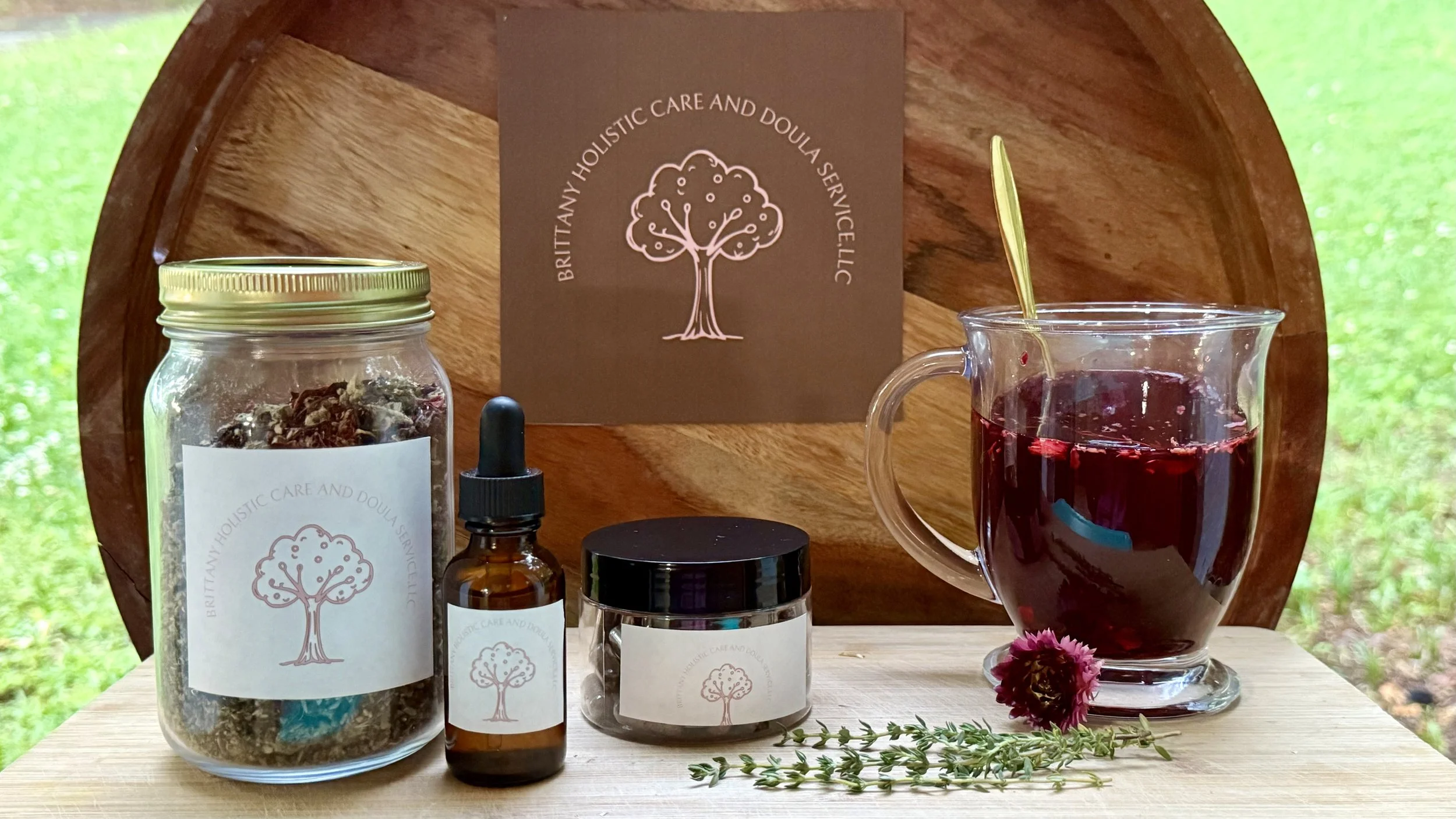 A wooden tray displaying various herbal products including dried herbs in a jar, small bottles, and jars labeled with a tree logo from Britanny Holistic Care and Doula Service. There is a glass pitcher of dark herbal tea, and fresh herbs and a pink flower are placed on the table in front.